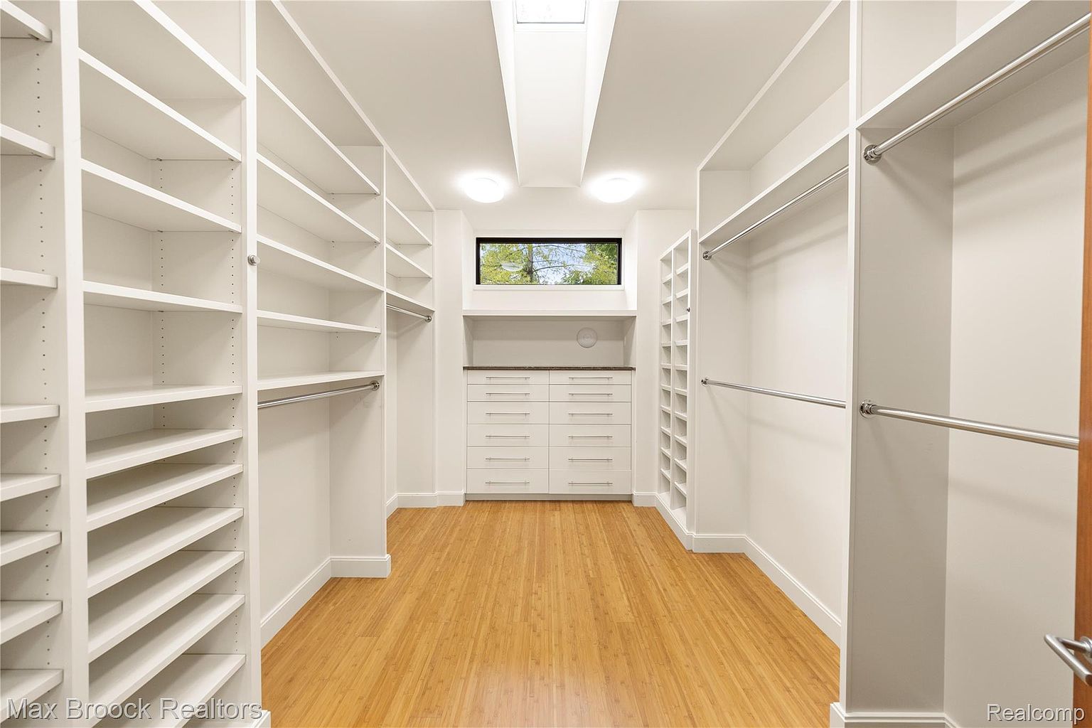 This is a well-organized walk-in closet featuring custom white shelving and drawers. The closet is brightly lit with a window and overhead lighting, and the hardwood floors add a touch of warmth. The space is designed for optimal storage and organization.