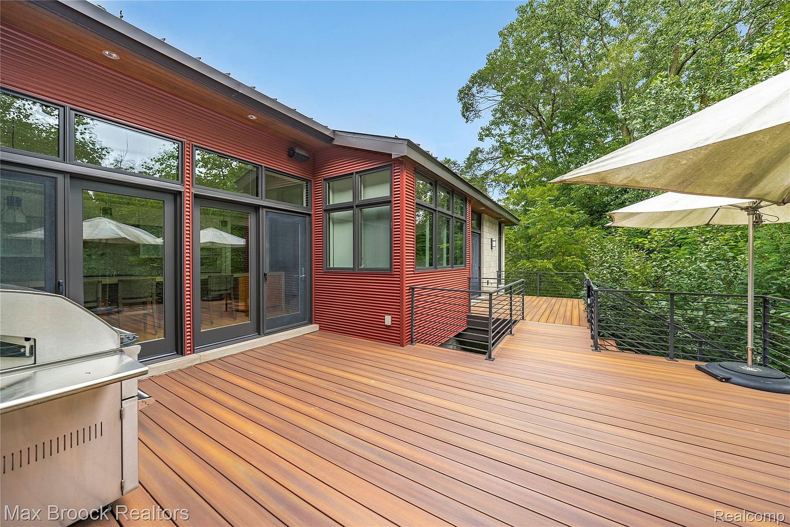 This image showcases a modern home's expansive wooden deck, featuring a sleek design with horizontal railings and a contemporary outdoor umbrella. The exterior of the house is clad in a striking red corrugated material, complemented by large windows and doors that offer seamless indoor-outdoor living. A stainless steel grill is visible, suggesting an ideal space for outdoor entertaining and relaxation.