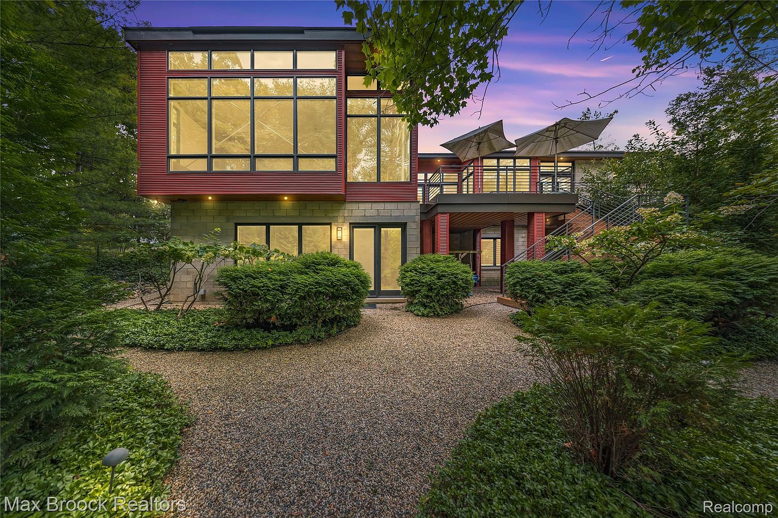 This is a front exterior view of a modern two-story home. The house features a combination of concrete block and red siding, with large windows providing ample natural light. The landscaping includes gravel pathways and lush greenery, creating a welcoming approach to the property.