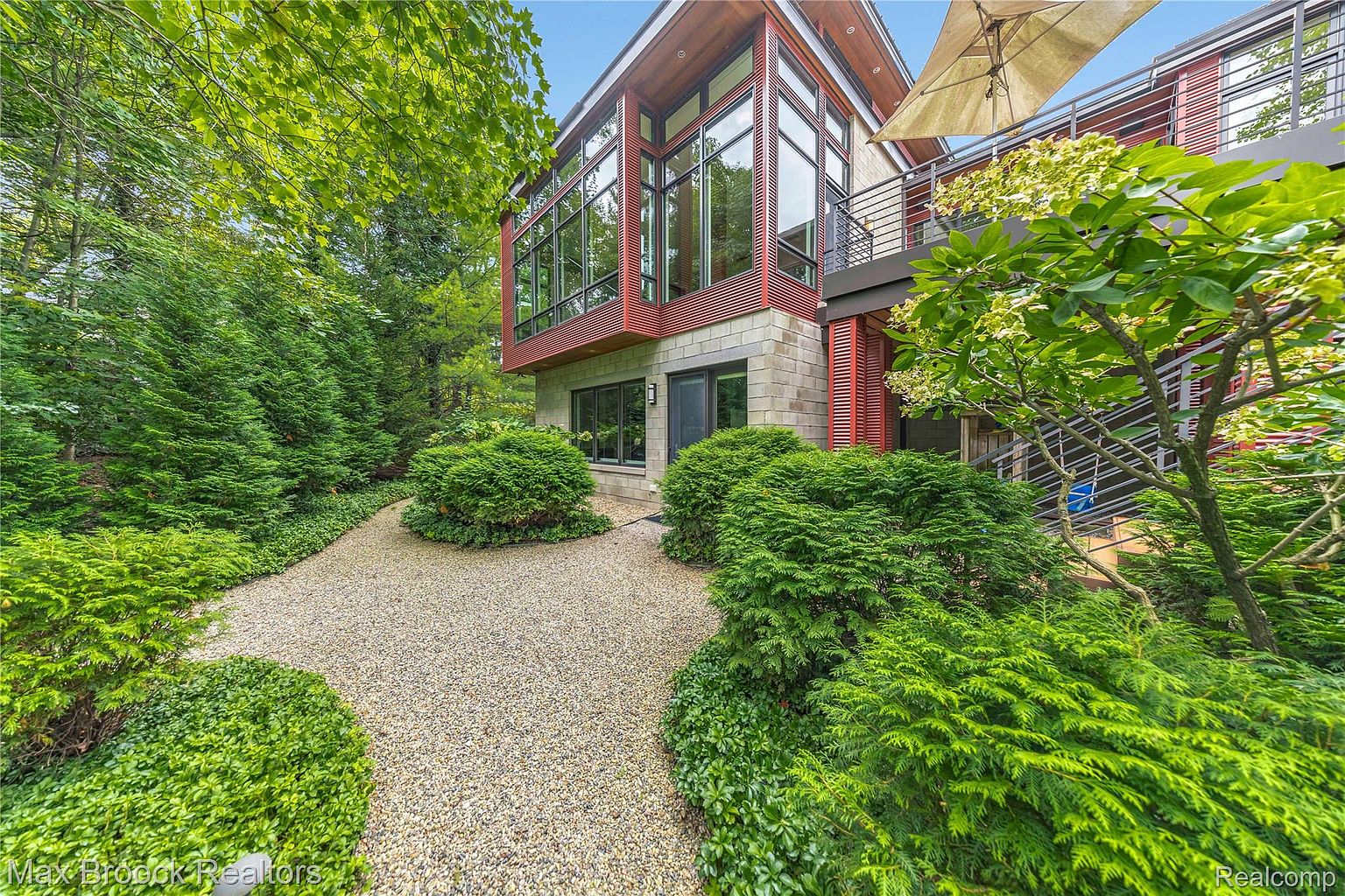 This image showcases the front exterior of a modern home with a unique architectural design. The building features a combination of stone and wood accents, large windows, and a well-maintained gravel pathway leading to the entrance. Lush greenery and landscaping surround the property, enhancing its curb appeal and creating a serene atmosphere.