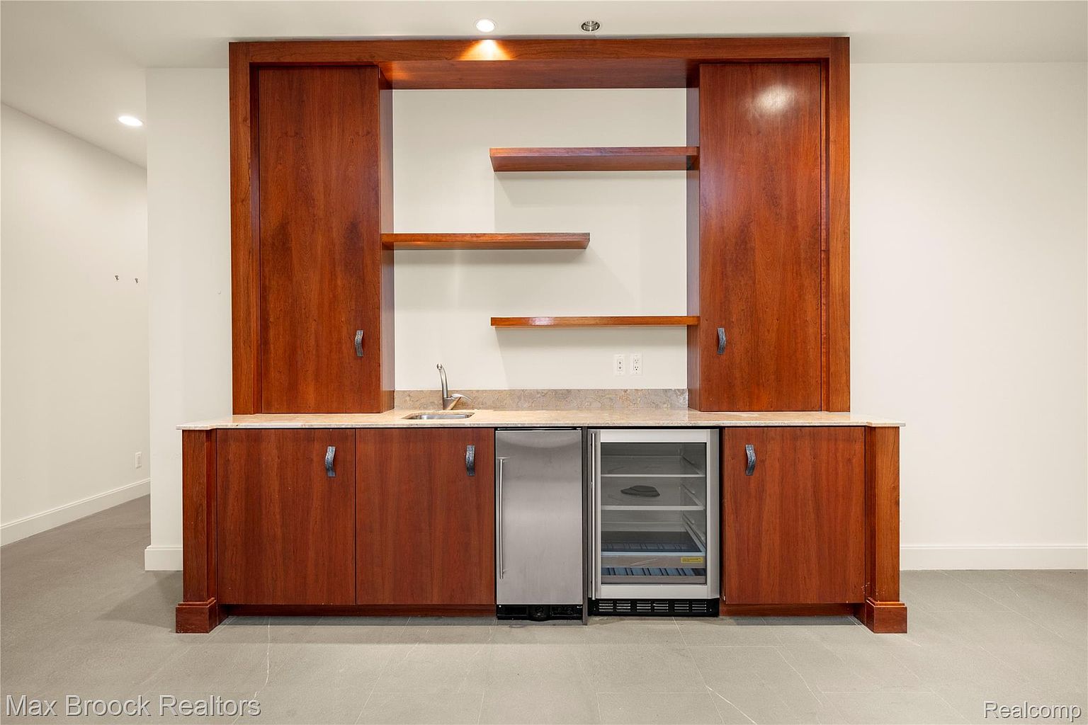 The image showcases a well-appointed basement bar area, featuring rich wood cabinetry with dark hardware, a marble countertop, and a stainless steel beverage refrigerator. Open shelving and recessed lighting add to the ambiance, while the gray tile flooring provides a modern touch. The perspective is a medium shot, highlighting the bar's features.