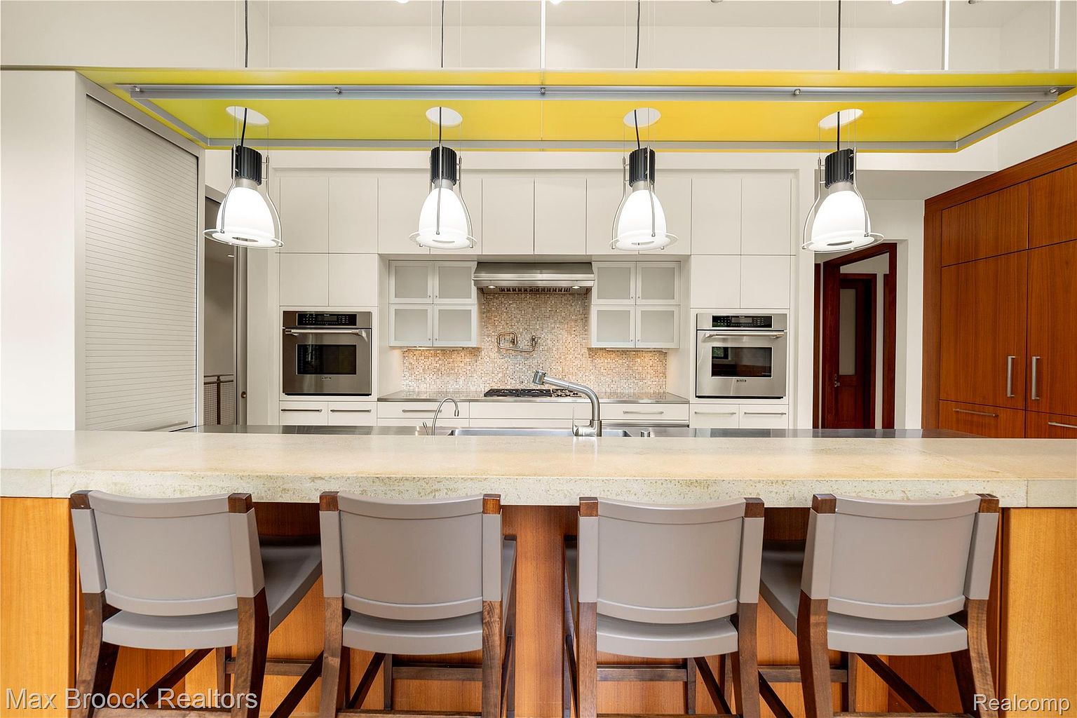 This is a well-lit kitchen featuring a large island with seating, white cabinetry, and stainless steel appliances. The kitchen has a modern design with a yellow accent above the cabinets and pendant lighting over the island. The perspective is from behind the island, showcasing the seating and the kitchen layout.