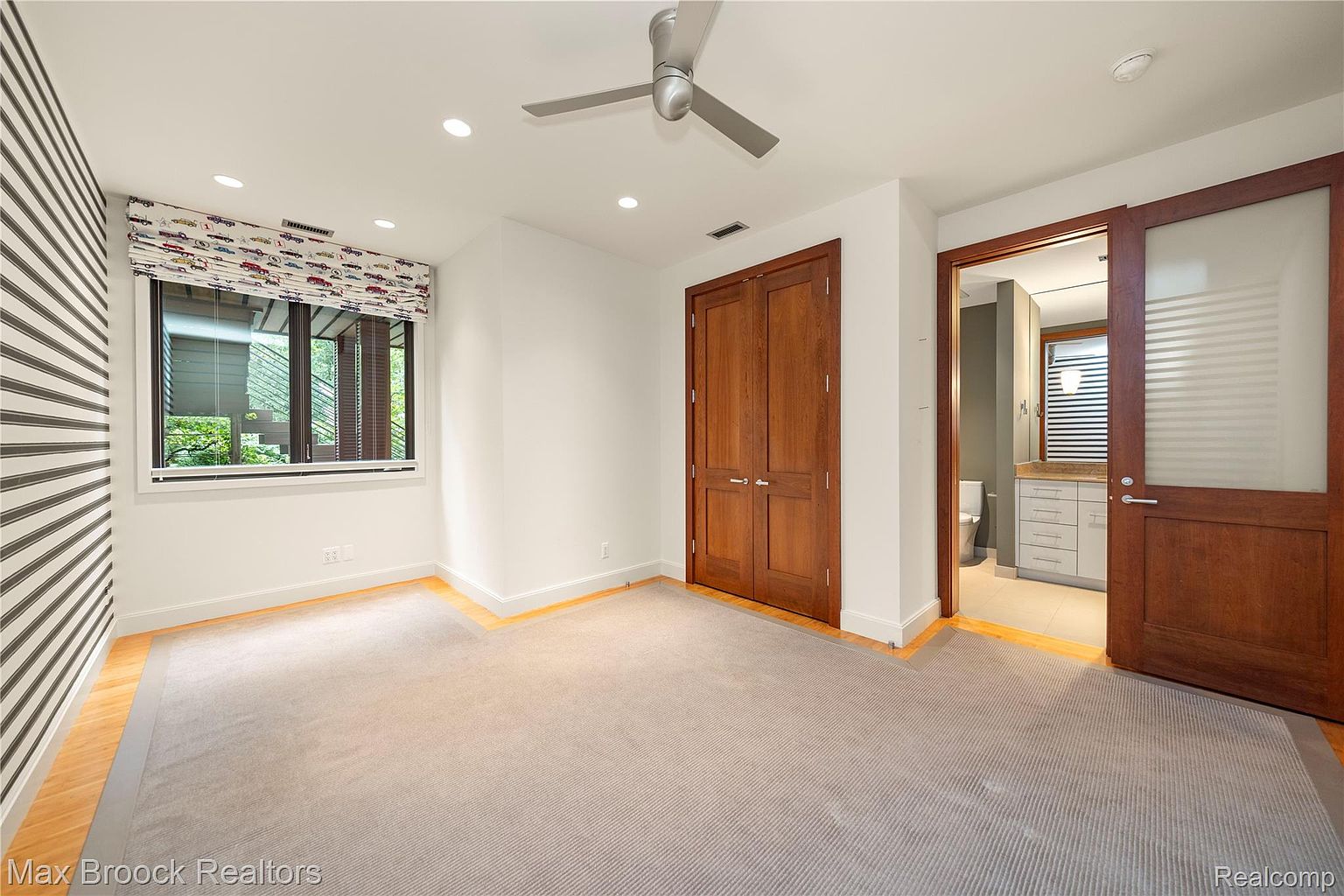 This is a well-lit bedroom featuring a neutral color palette with a striped accent wall and a window with patterned blinds. The room includes a double-door closet and an open doorway leading to a bathroom, enhancing the sense of space and convenience. The overall impression is clean and inviting, suitable for a guest or family member.