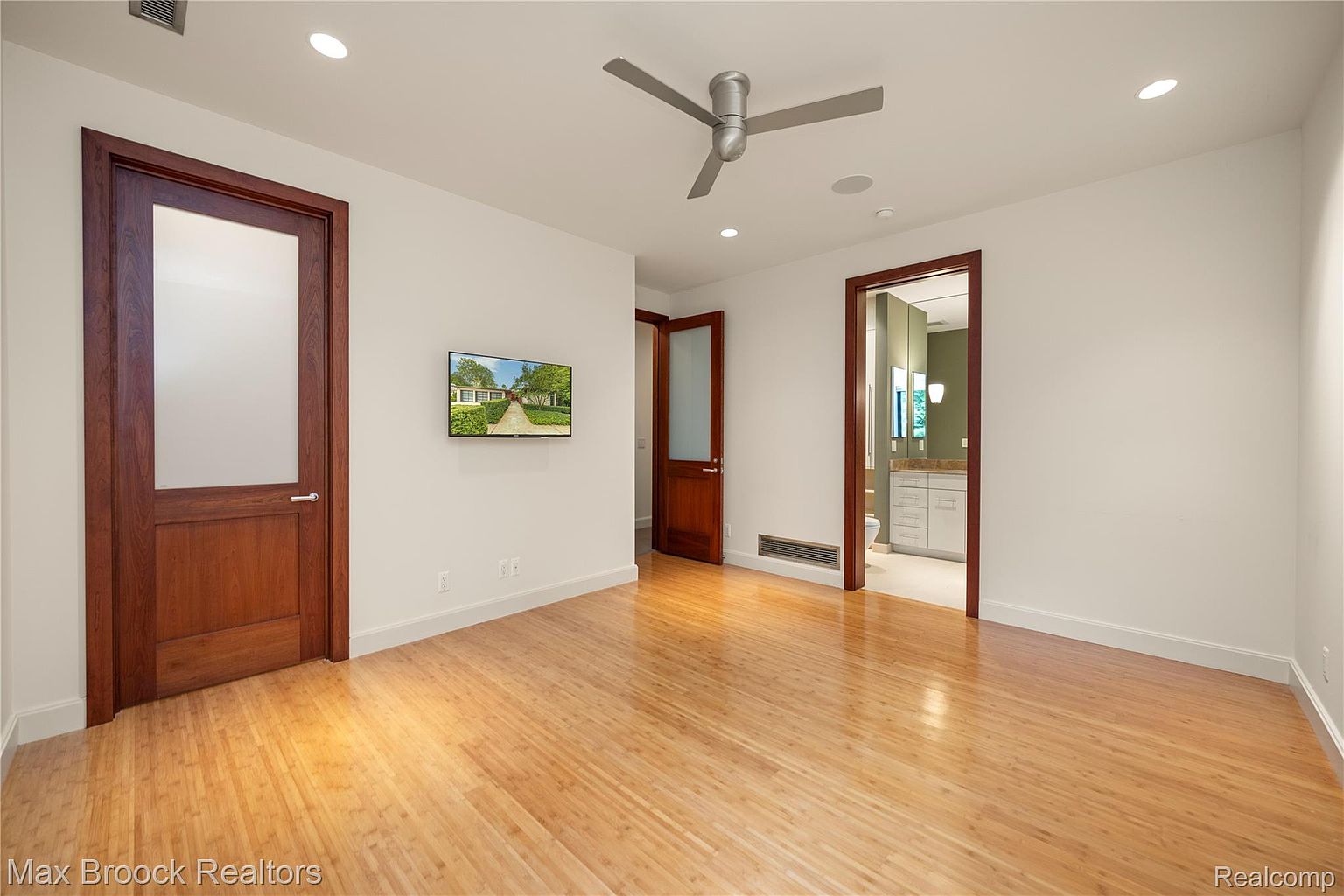 This is a spacious primary bedroom featuring hardwood floors and neutral wall colors. The room has multiple doorways, including one leading to a bathroom, and a ceiling fan. A flat-screen television is mounted on the wall, and the overall impression is clean and modern.