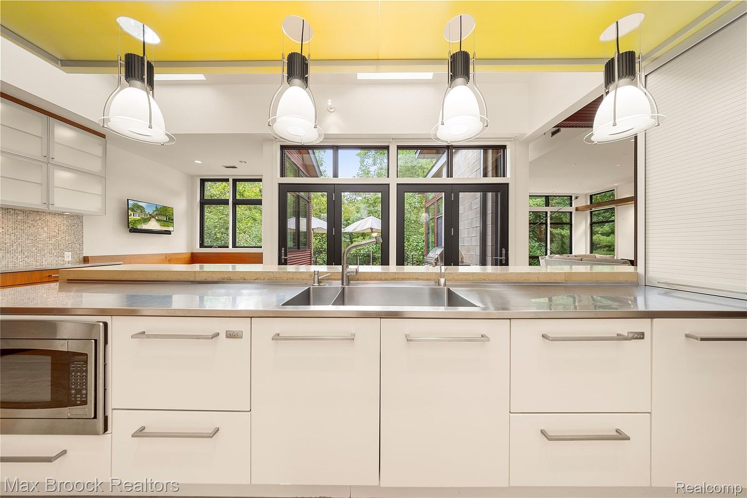 This is a modern kitchen with sleek white cabinetry and stainless steel countertops. The kitchen features a double sink, stainless steel appliances, and unique pendant lighting. Large windows provide natural light and a view to the outside, enhancing the bright and airy feel of the space.