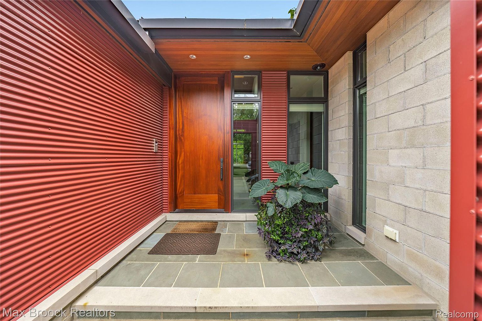 This image showcases a modern home's entryway, featuring a striking red corrugated metal wall contrasted by a light-colored brick section. A solid wood door is flanked by narrow windows, and a large potted plant adds a touch of greenery. The entryway is paved with large, gray stone tiles, creating a clean and inviting aesthetic.