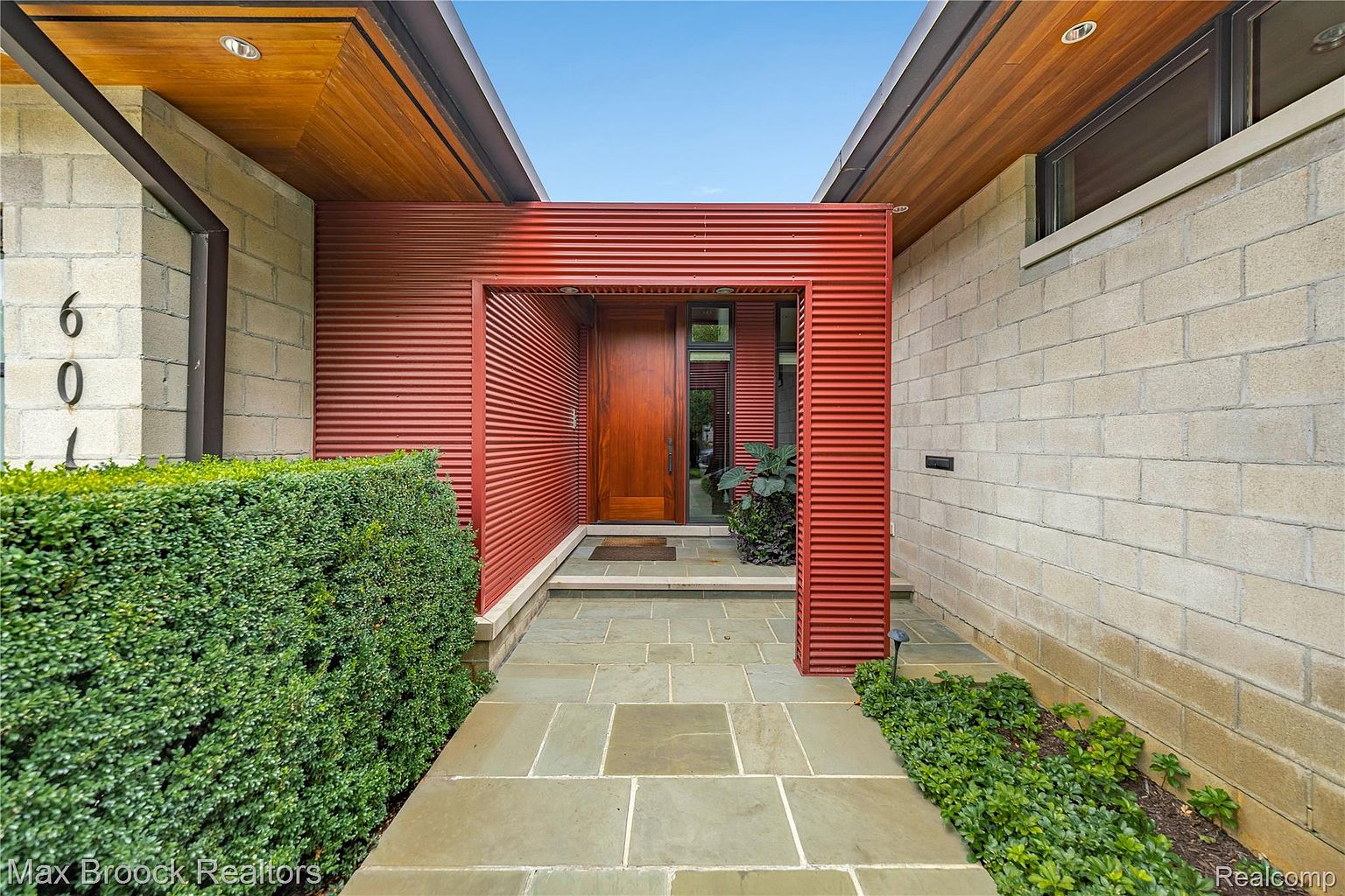 This is a front entryway of a modern home, featuring a red corrugated metal accent wall framing a wooden front door. The walkway is paved with large square stones, flanked by manicured greenery. The house's exterior is constructed with light-colored concrete blocks, and the overall design is clean and contemporary.