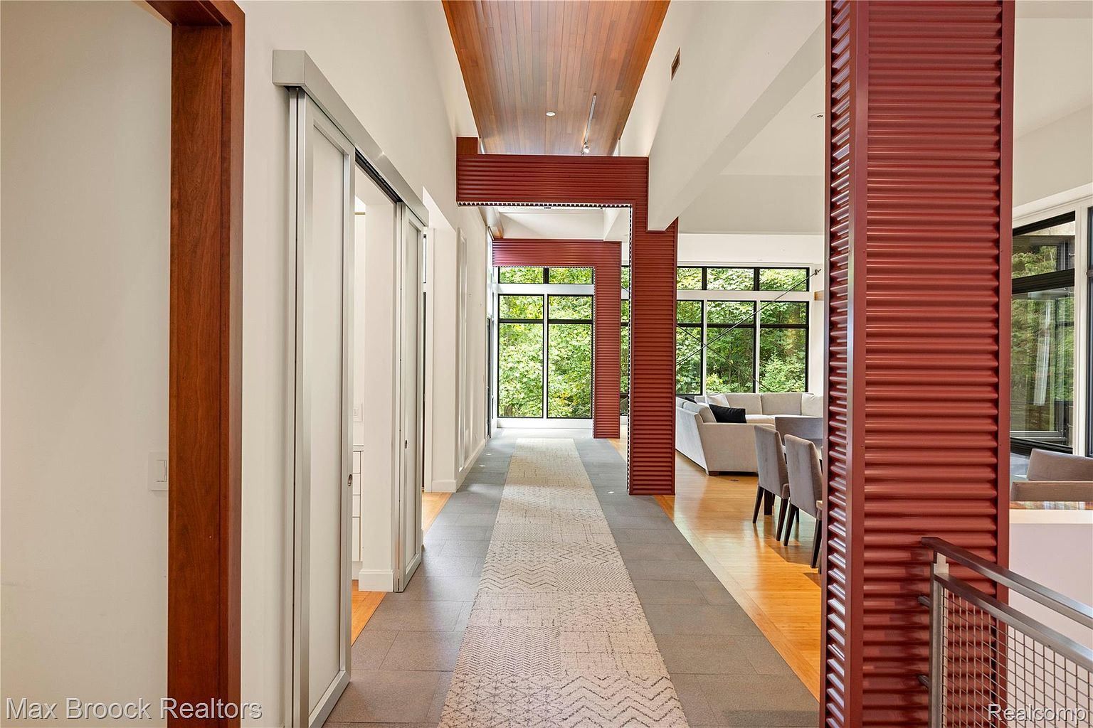 This interior shot showcases a modern hallway with a unique architectural design. The hallway features a combination of tile and carpet flooring, with sliding doors on one side and red-toned structural columns that partially obscure the view into a living area with large windows. The ceiling has a mix of white and wood paneling, creating a visually interesting space.