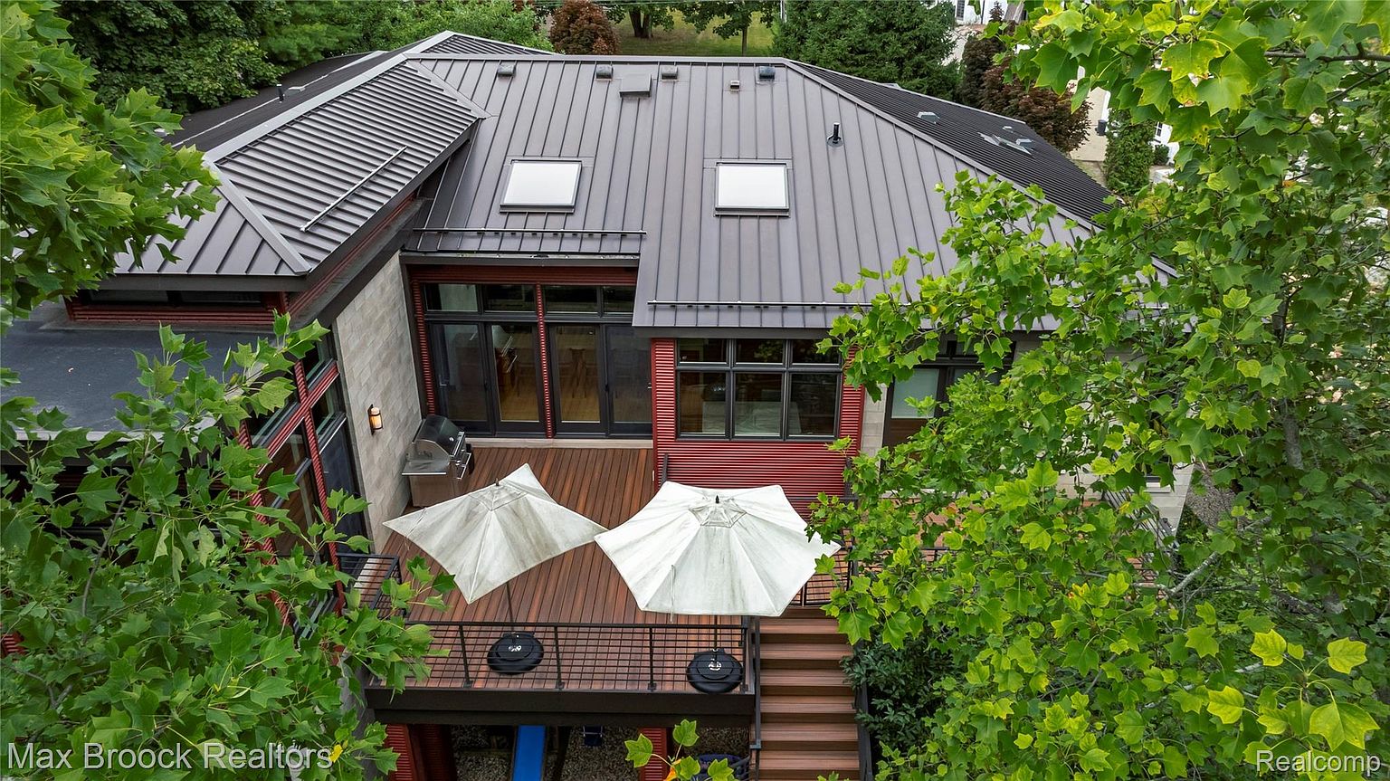 This aerial view showcases the rear of a modern home with a striking metal roof and a spacious wooden deck. The deck features outdoor seating areas with umbrellas, and the home's exterior combines stone, red siding, and large windows. Lush greenery surrounds the property, providing privacy and a natural aesthetic.