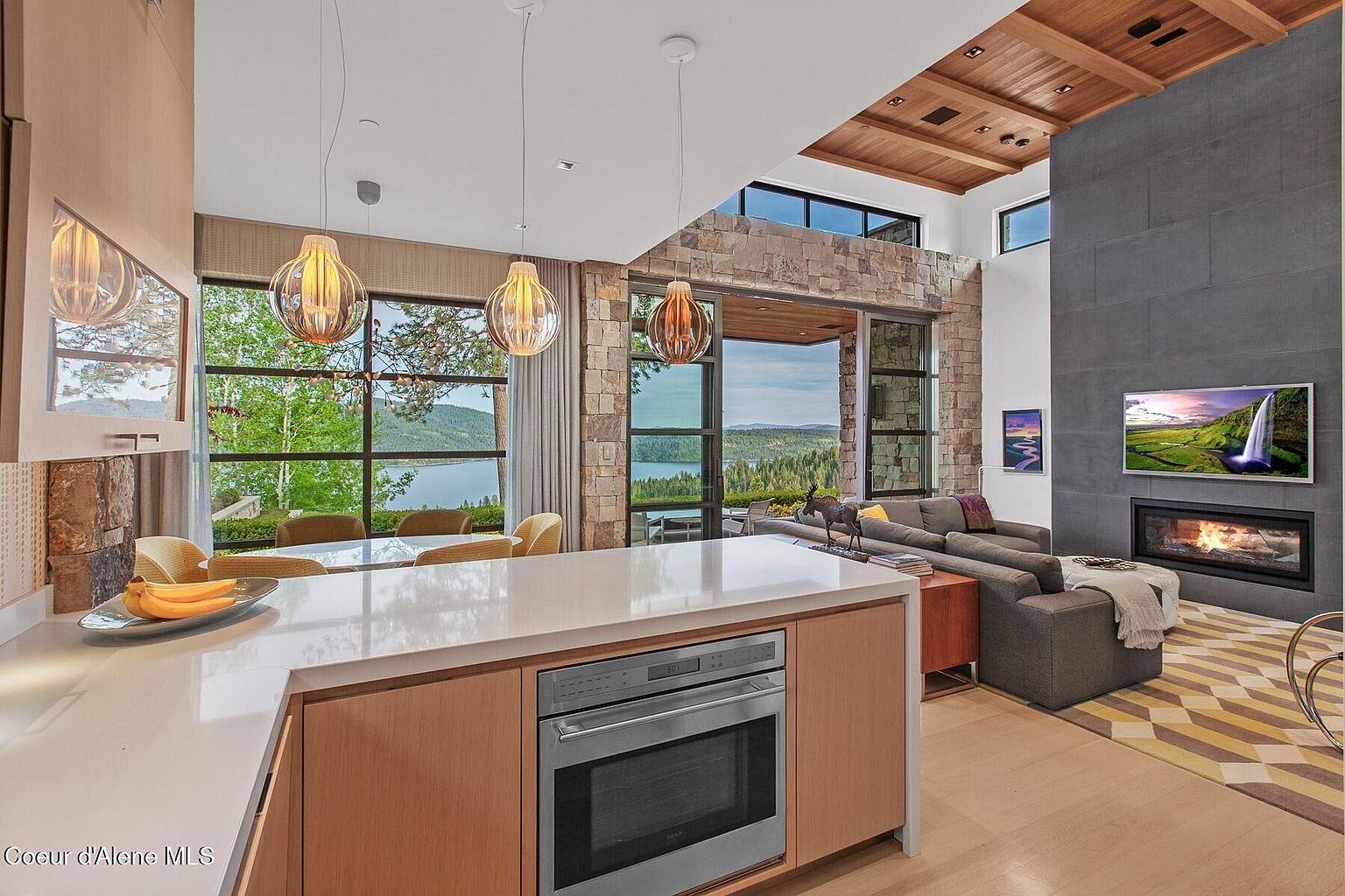 This interior shot showcases a modern kitchen seamlessly integrated with a living area. The kitchen features a sleek island with a built-in oven and a white countertop, complemented by warm wood cabinetry. Large windows offer stunning views of a lake and surrounding landscape, while the living area boasts a stone fireplace, a comfortable gray sofa, and a geometrically patterned rug, creating a luxurious and inviting atmosphere.