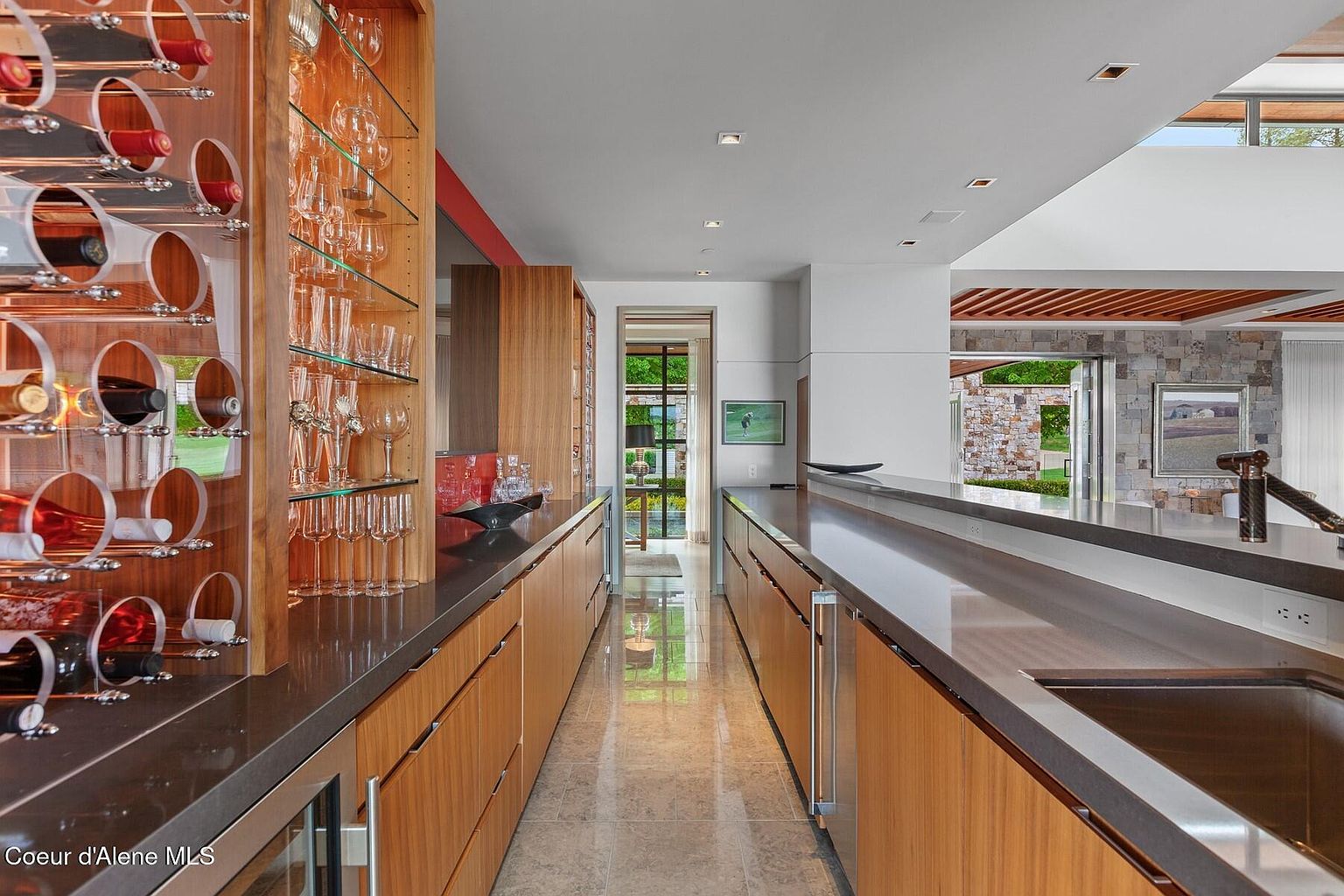 This interior shot showcases a modern wine cellar and bar area. The space features custom wooden cabinetry with built-in wine racks and glass displays for stemware. A sleek countertop runs along both sides of the hallway, creating a sophisticated and functional space for wine storage and service.
