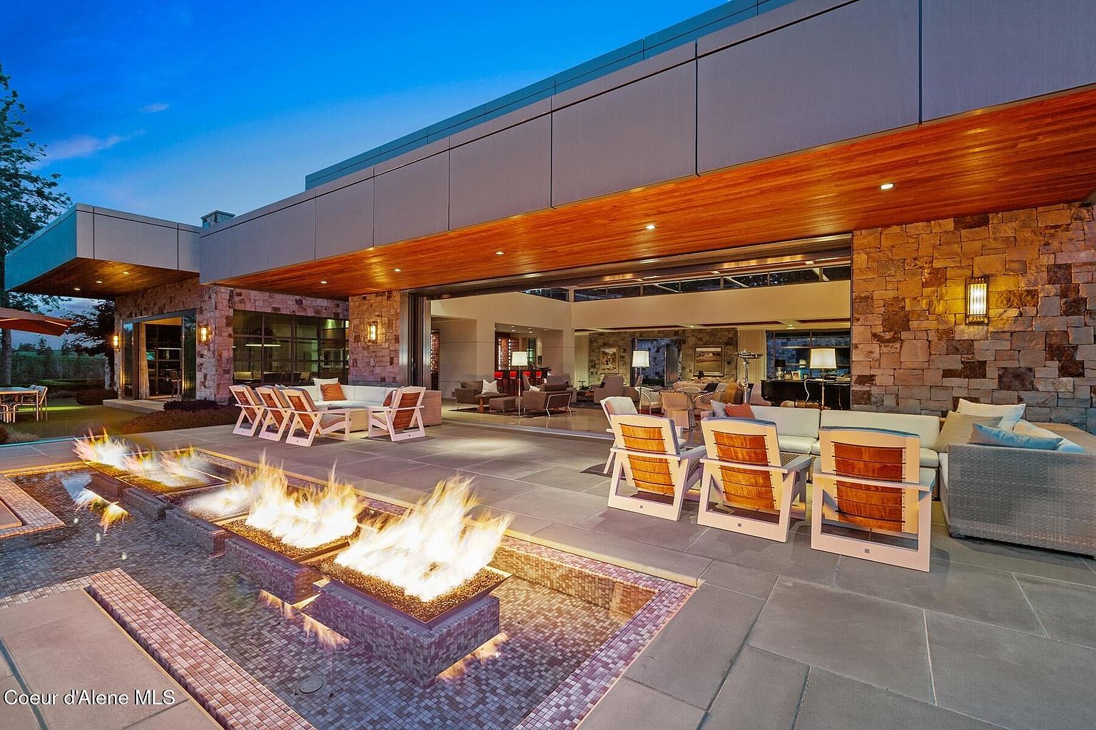 This image showcases a luxurious outdoor patio area featuring a modern fire pit with flames, comfortable seating arrangements, and a seamless transition to the interior through large sliding glass doors. The patio is constructed with stone and tile, complemented by a stone exterior wall and wood ceiling, creating an inviting and sophisticated outdoor living space. The open layout and elegant design make it perfect for entertaining or relaxing.