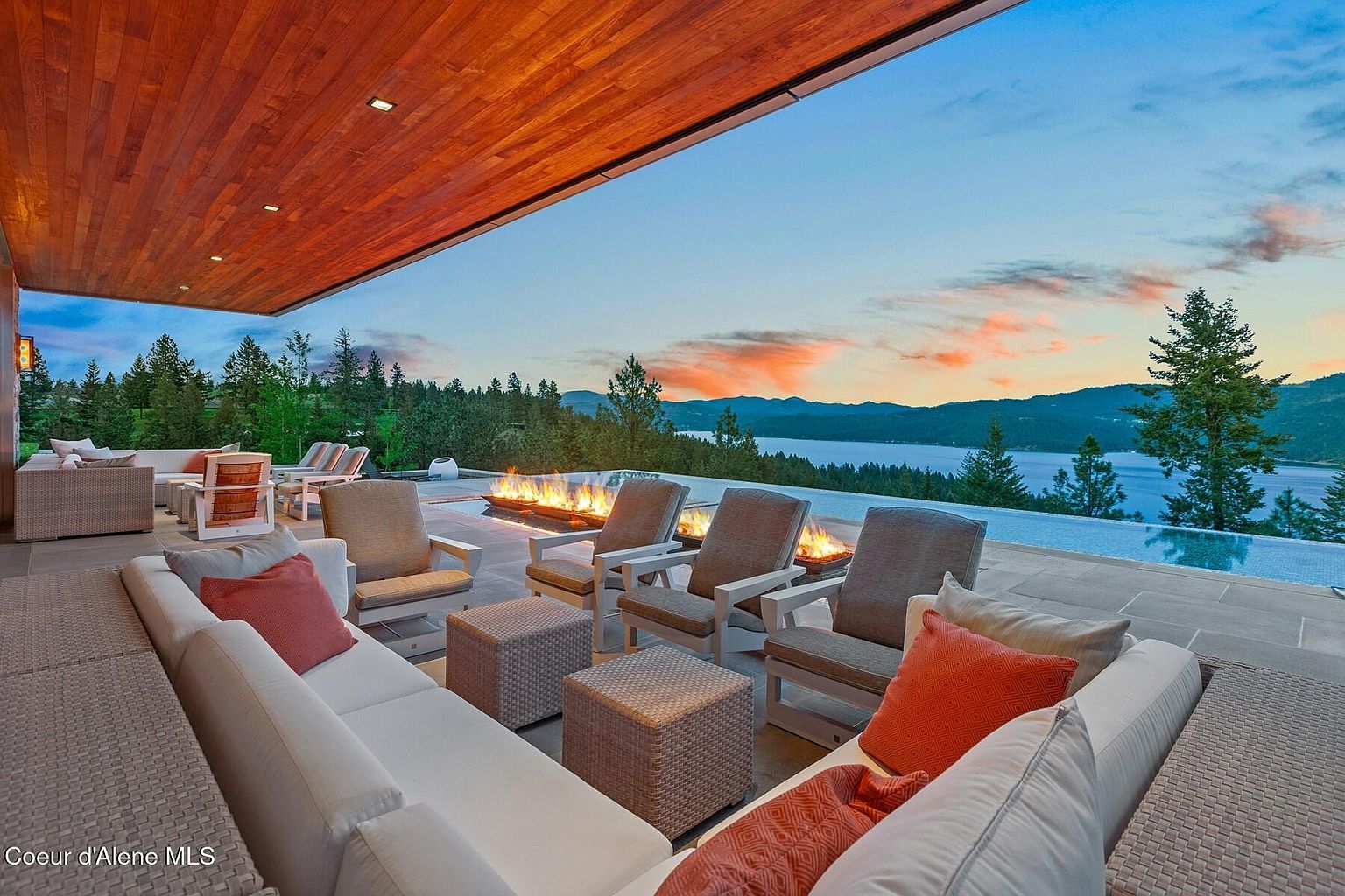 This image showcases a luxurious outdoor patio area with comfortable seating, including a large sectional sofa with decorative pillows and several armchairs. A modern fire pit adds warmth and ambiance, while an infinity pool offers stunning views of the lake and surrounding landscape at sunset. The wooden ceiling adds a touch of elegance to the outdoor space.
