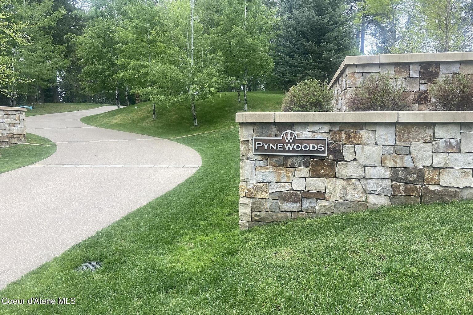 The image showcases the entryway to a community named "PyneWoods", marked by a stone wall with a sign. A curved paved driveway leads into the community, bordered by lush green lawns and mature trees. The scene conveys a sense of upscale residential living and well-maintained landscaping.