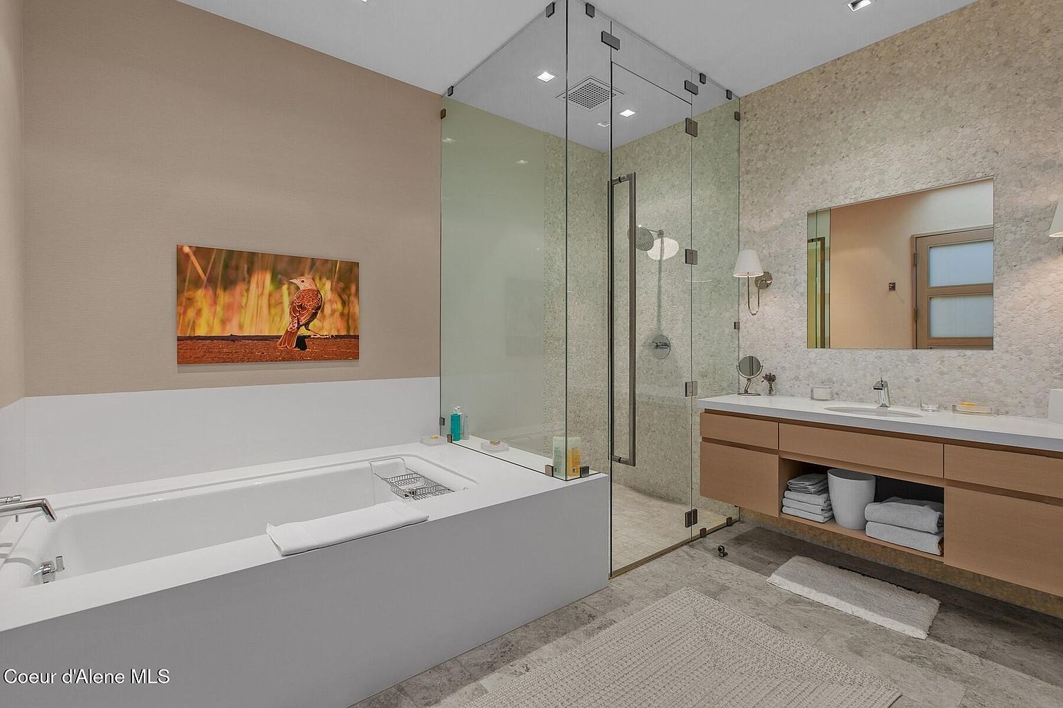 This is a luxurious primary bathroom featuring a modern freestanding bathtub, a glass-enclosed shower with rainfall showerhead, and a sleek vanity with ample storage. The walls are adorned with textured tile, and a large mirror reflects natural light, creating a spa-like atmosphere. The overall design is contemporary and elegant.