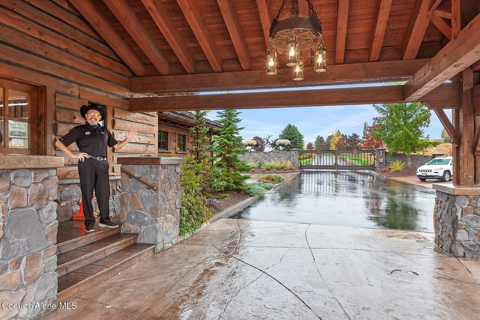 This image showcases the grand entryway of a luxurious property, featuring a covered porte-cochère with exposed wooden beams and stone pillars. The driveway leads to an ornate gate with elk statues, and a white SUV is parked nearby. The overall impression is one of upscale elegance and privacy.