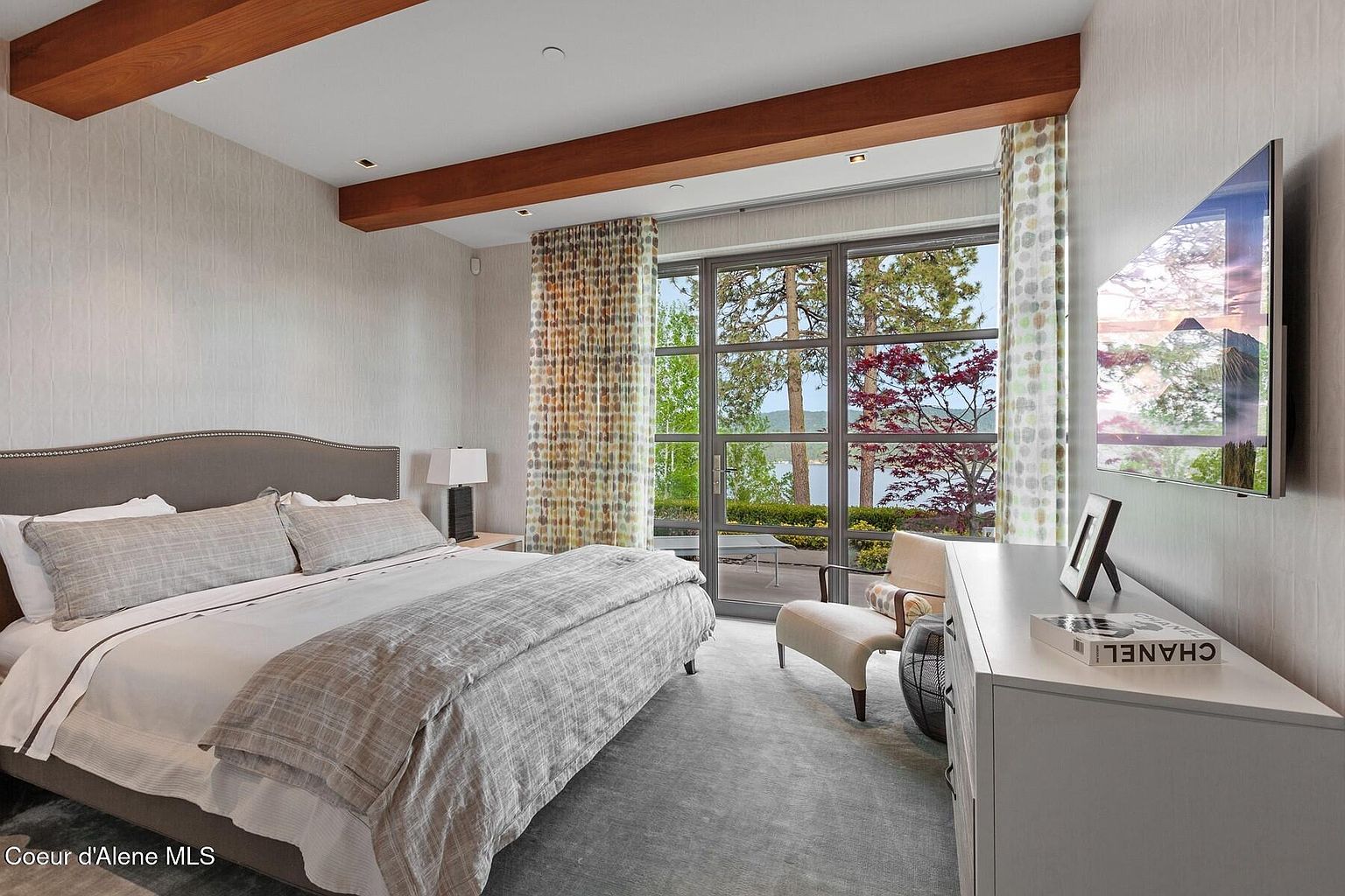 This is a primary bedroom featuring a large bed with neutral-toned bedding, wooden ceiling beams, and a wall-mounted television. A large window offers a view of the outdoors, and a chair with ottoman sits near the window. The room has a calm and luxurious feel.