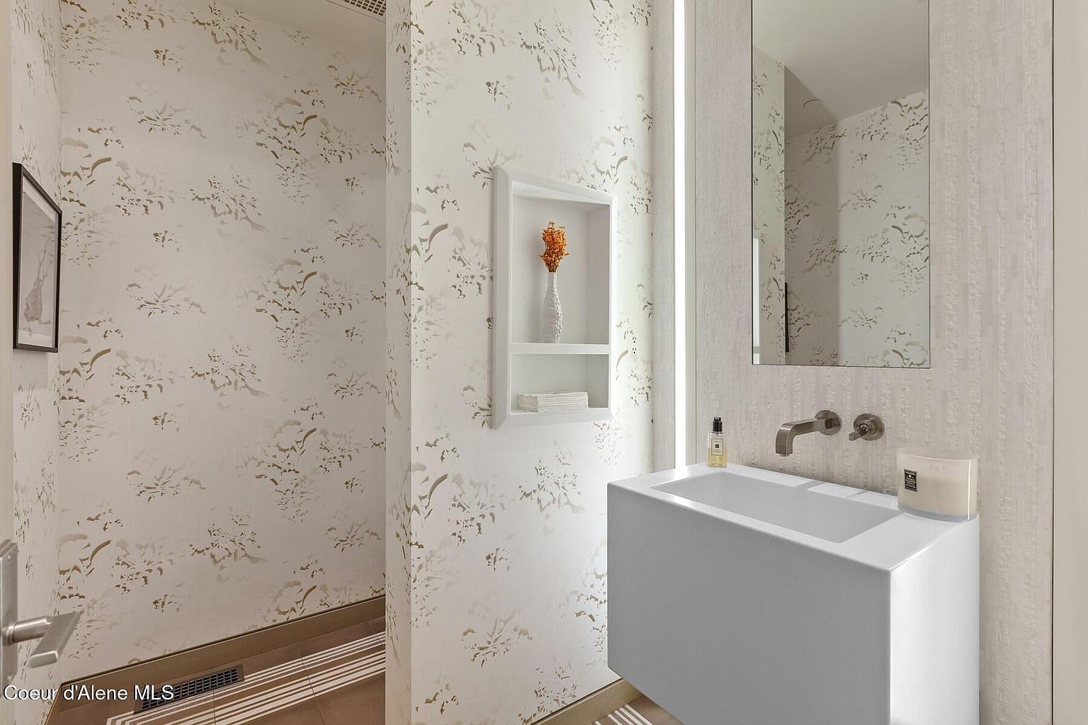 This is a stylish guest bathroom featuring a modern, wall-mounted sink with a sleek faucet and a rectangular mirror. The walls are adorned with a subtle floral wallpaper, complemented by a built-in niche displaying decorative items. The overall aesthetic is clean, contemporary, and inviting, creating a sophisticated space for guests.