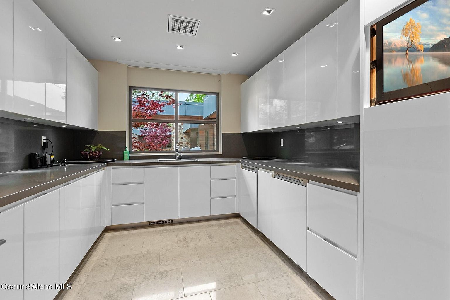 This is a modern kitchen with sleek, white cabinetry and dark countertops. A large window over the sink provides natural light and a view of the outdoors. The kitchen features a clean, minimalist design with a built-in television on the right side.