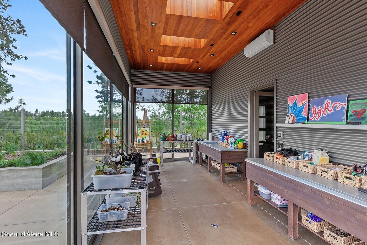 This sunroom features a wood plank ceiling with skylights and corrugated metal walls. Large windows offer views of the surrounding landscape. The room is furnished with work tables and shelving, suggesting a space for hobbies or gardening.