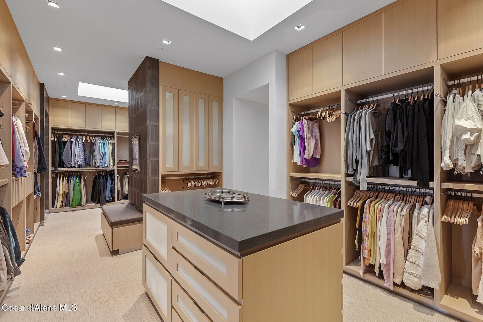 This is a well-organized walk-in closet featuring custom cabinetry and ample storage space for clothing and accessories. The closet includes hanging rods, drawers, and shelving, all illuminated by natural light from a skylight. A central island with a dark countertop provides additional surface area and storage.