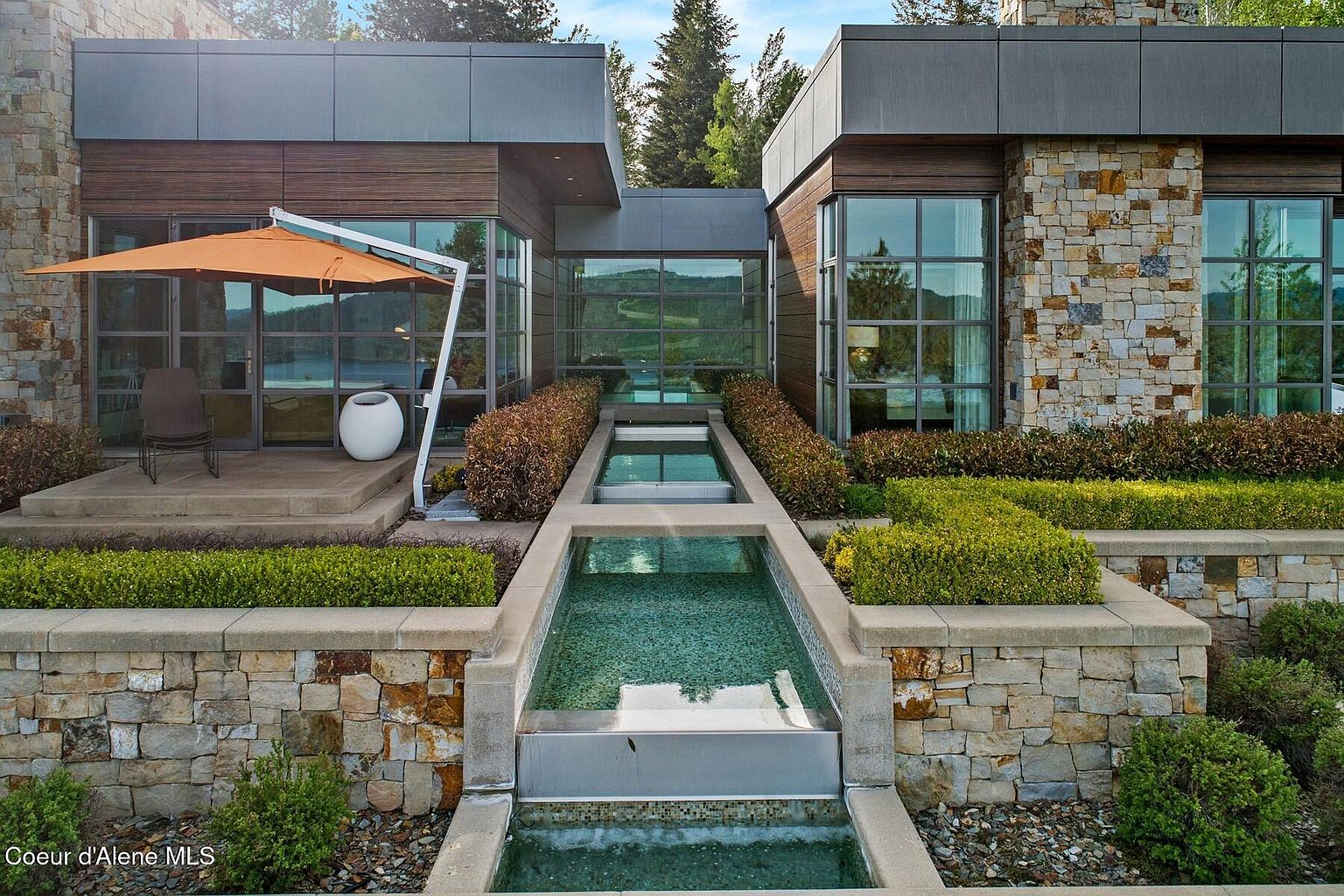This is a front exterior view of a modern home featuring a unique water feature leading to the entrance. The architecture combines stone and wood elements, with large windows providing ample natural light. The landscaping is meticulously maintained, adding to the property's curb appeal.