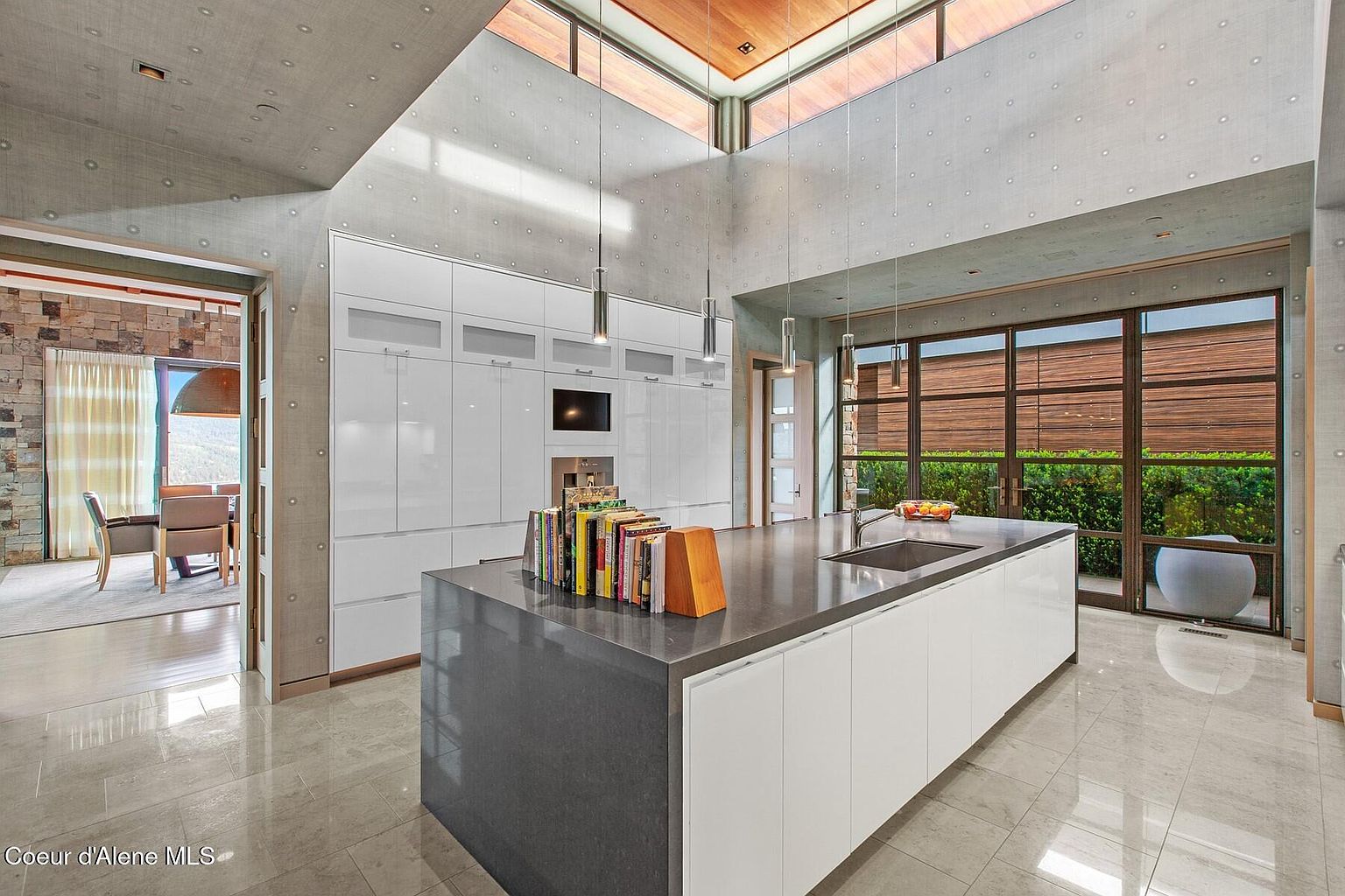 This is a modern kitchen with a large island featuring a gray countertop and white cabinetry. The kitchen has high ceilings with skylights and pendant lighting. A wall of white cabinets houses appliances, and a large window offers a view of the greenery outside, creating a bright and airy atmosphere.