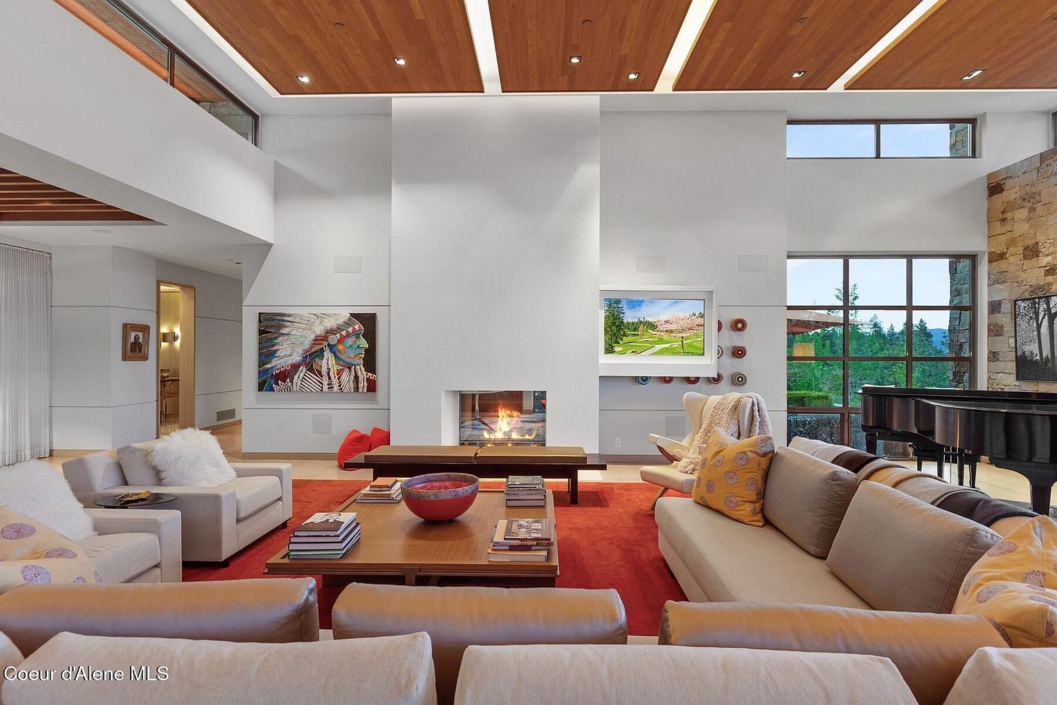 This is a spacious living room featuring a modern design with a high ceiling and wooden accents. The room is furnished with multiple sofas, a coffee table with books, and a fireplace. Large windows provide natural light and views of the outdoors, while artwork adds a touch of personality to the space.