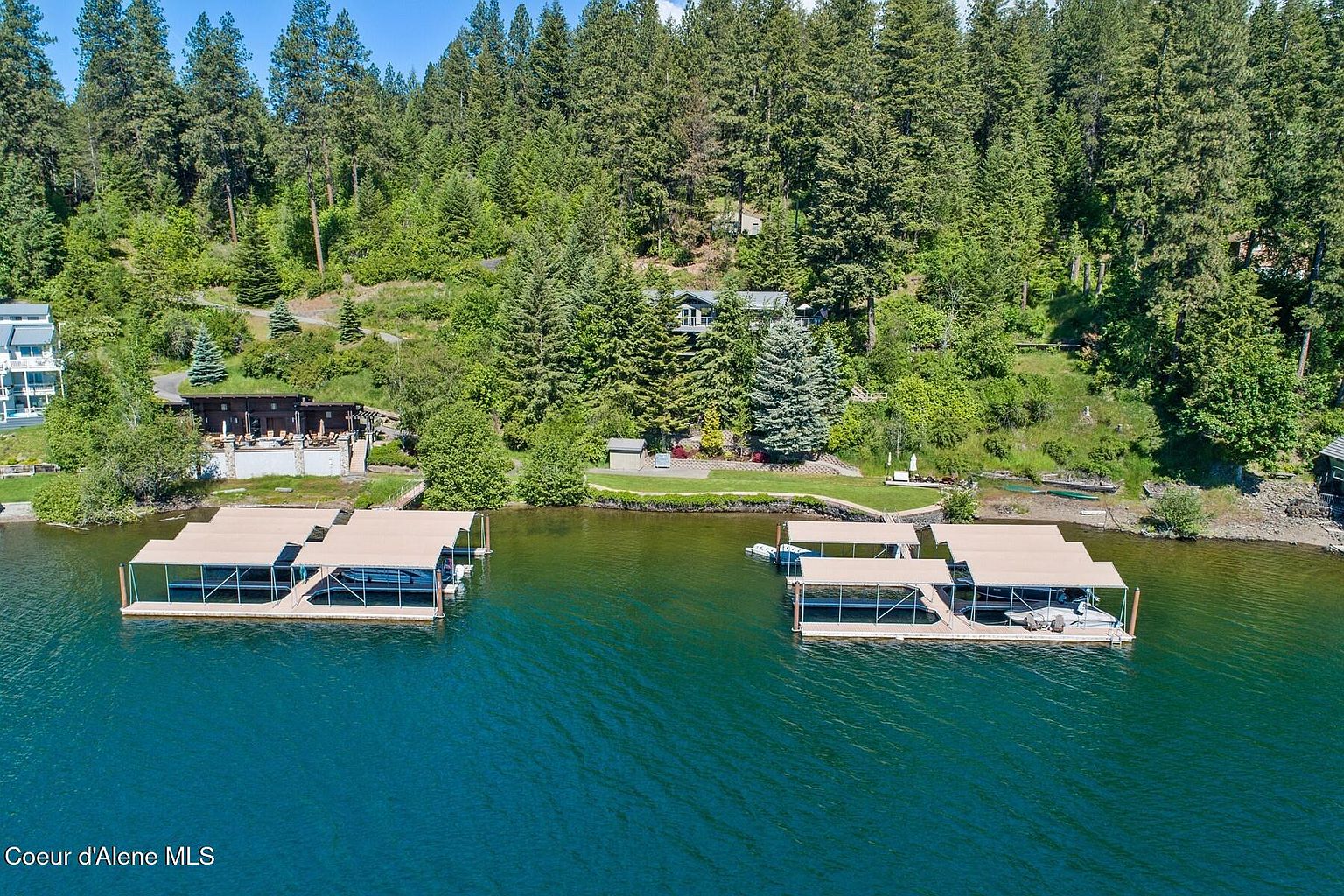 This aerial shot showcases a waterfront property with two large docks featuring boat covers on a serene lake surrounded by lush, green forest. The homes on the property are nestled among the trees, offering privacy and scenic views. The overall impression is one of luxury and tranquility, ideal for a lakeside retreat.