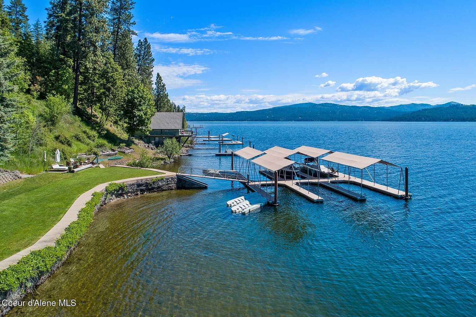 This aerial view showcases a stunning waterfront property featuring a well-maintained lawn, a charming house nestled among trees, and a substantial dock with multiple boat slips. The clear blue water and mountain backdrop enhance the property's appeal, creating a picturesque and desirable setting. The perspective is high and wide, capturing the scale and beauty of the estate.