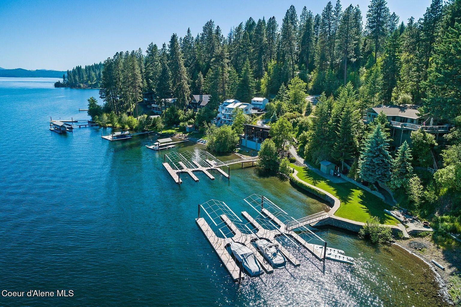 This aerial view showcases a stunning waterfront property with multiple docks and boat slips. The property is nestled among lush, mature trees, offering privacy and a serene setting. Several houses are visible, blending into the natural landscape, and the clear blue water adds to the overall appeal of this lakeside retreat.