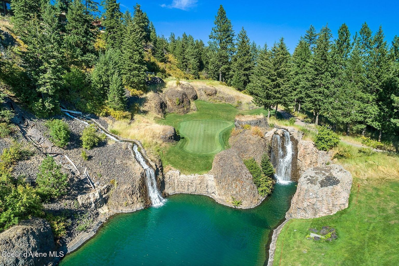 This aerial view showcases a stunning golf course feature with a green surrounded by rocky cliffs and waterfalls cascading into a turquoise pond. Lush trees frame the scene, creating a picturesque and luxurious setting. The image highlights the unique and desirable landscape of the property.
