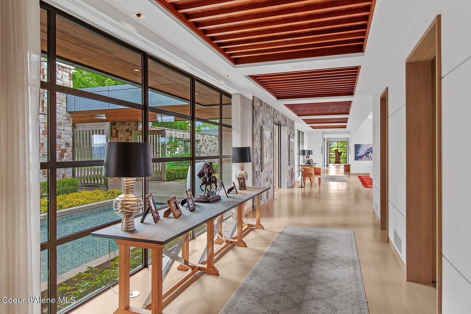 This interior shot showcases a long, well-lit hallway featuring a unique ceiling design with wooden beams. Large windows offer views of an exterior water feature and landscaping, while a console table displays decorative items. The hallway's design blends modern and natural elements, creating a sophisticated and inviting atmosphere.