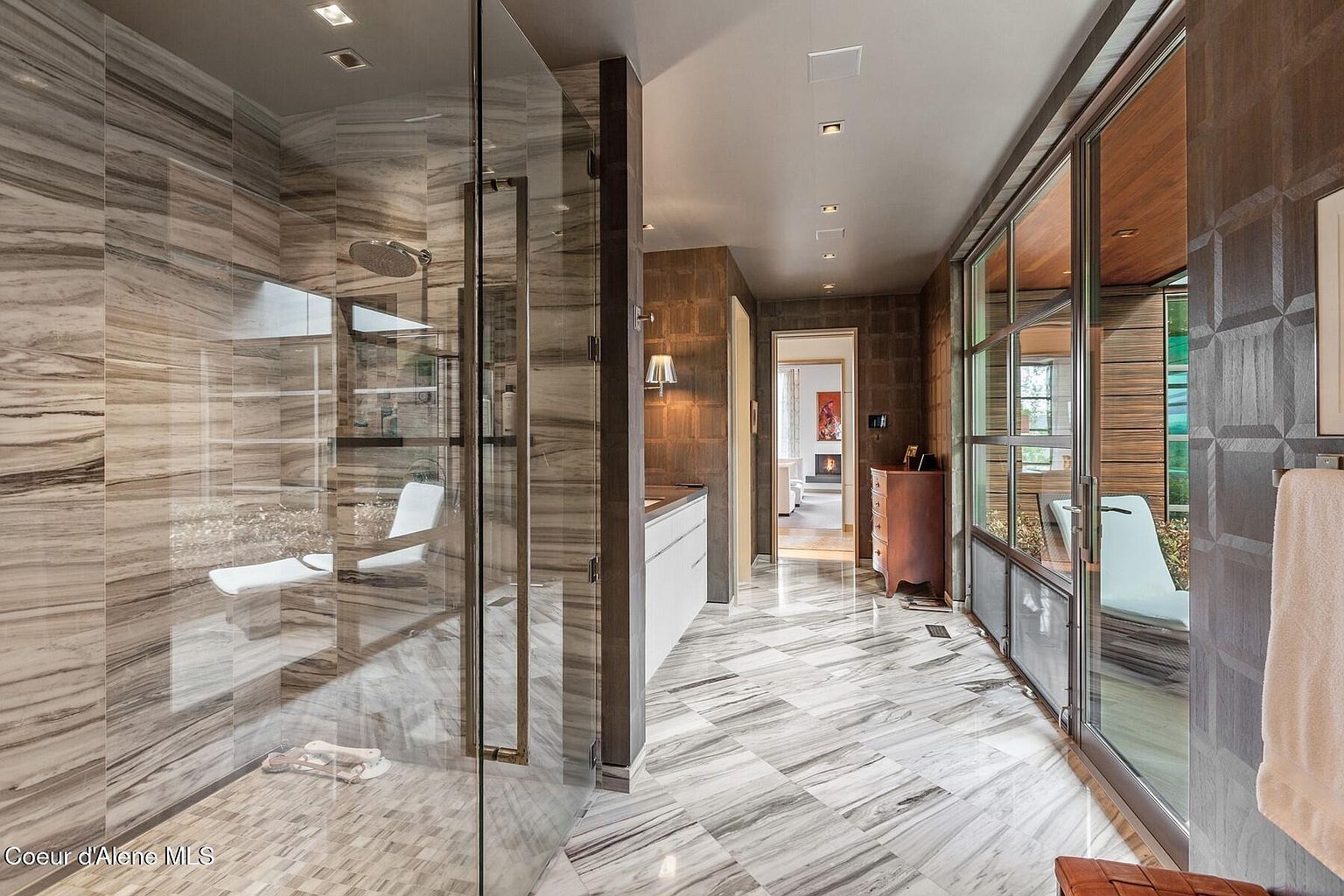 This is a luxurious primary bathroom featuring a spacious walk-in shower with marble walls and glass enclosure. The flooring is marble tile laid in a geometric pattern, and there's a modern vanity with sleek white cabinetry. A hallway leads to another room, and a glass-enclosed area with a lounge chair is visible, creating a spa-like atmosphere.