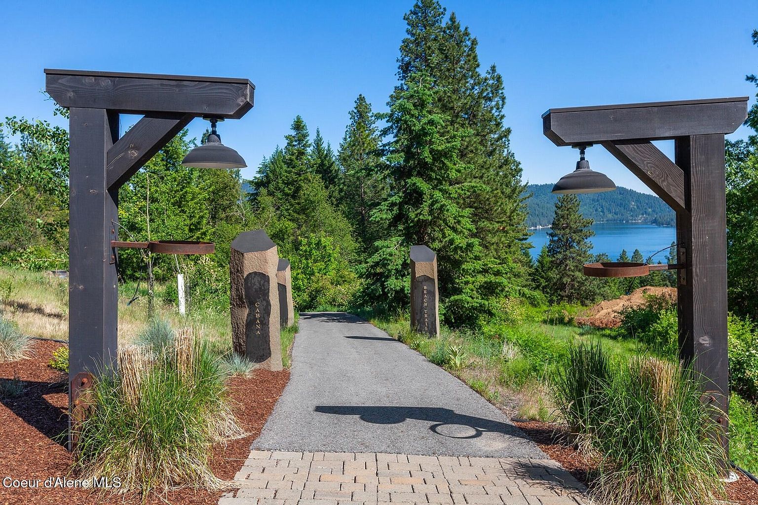 This image showcases an inviting entryway leading towards a property. The pathway is flanked by stone pillars with signage and wooden structures with hanging lamps, creating a sophisticated and welcoming ambiance. Lush greenery and a glimpse of a lake in the background add to the scenic appeal, suggesting a tranquil and upscale setting.