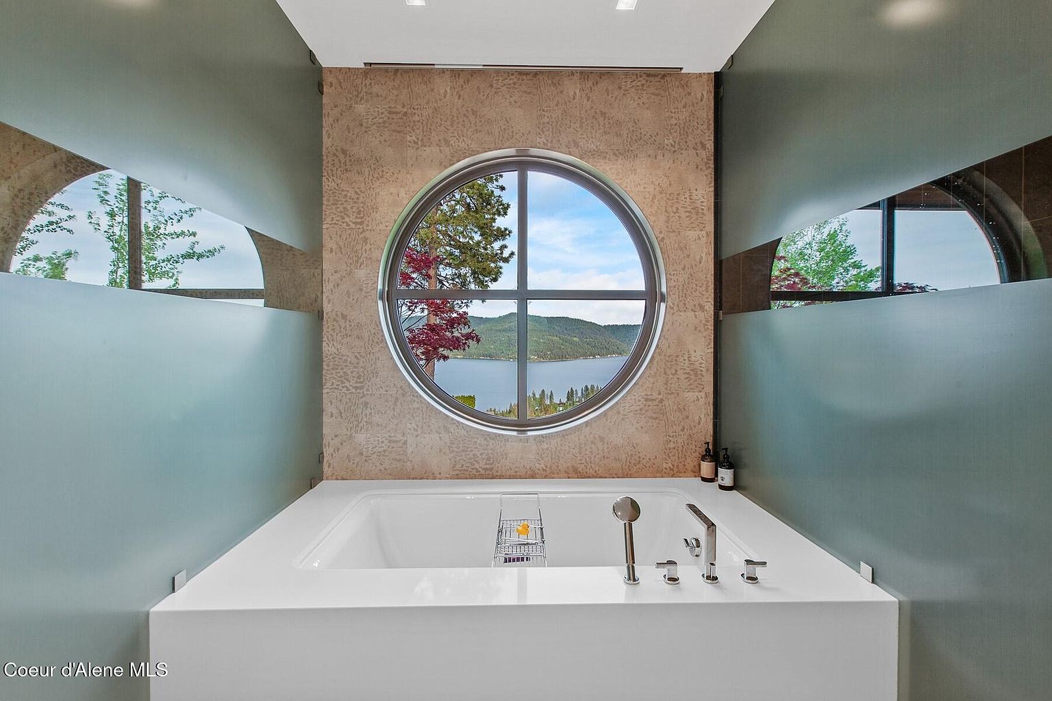 This is a luxurious primary bathroom featuring a modern, built-in bathtub with a sleek, white surround. A large, round window provides a stunning view of a lake and surrounding landscape, creating a serene and spa-like atmosphere. The walls are a combination of textured tile and muted green, adding to the room's sophisticated and calming aesthetic.