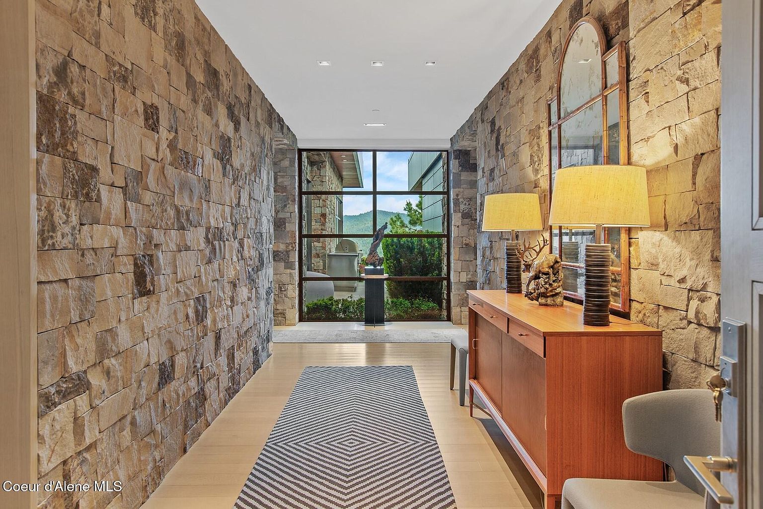 This interior shot showcases a hallway with stone walls and a light wood floor, creating a rustic yet modern aesthetic. A large window at the end of the hall provides natural light and a view of the outdoors, while a wooden console table with lamps and a mirror adds a touch of elegance. A patterned runner rug adds visual interest to the space.