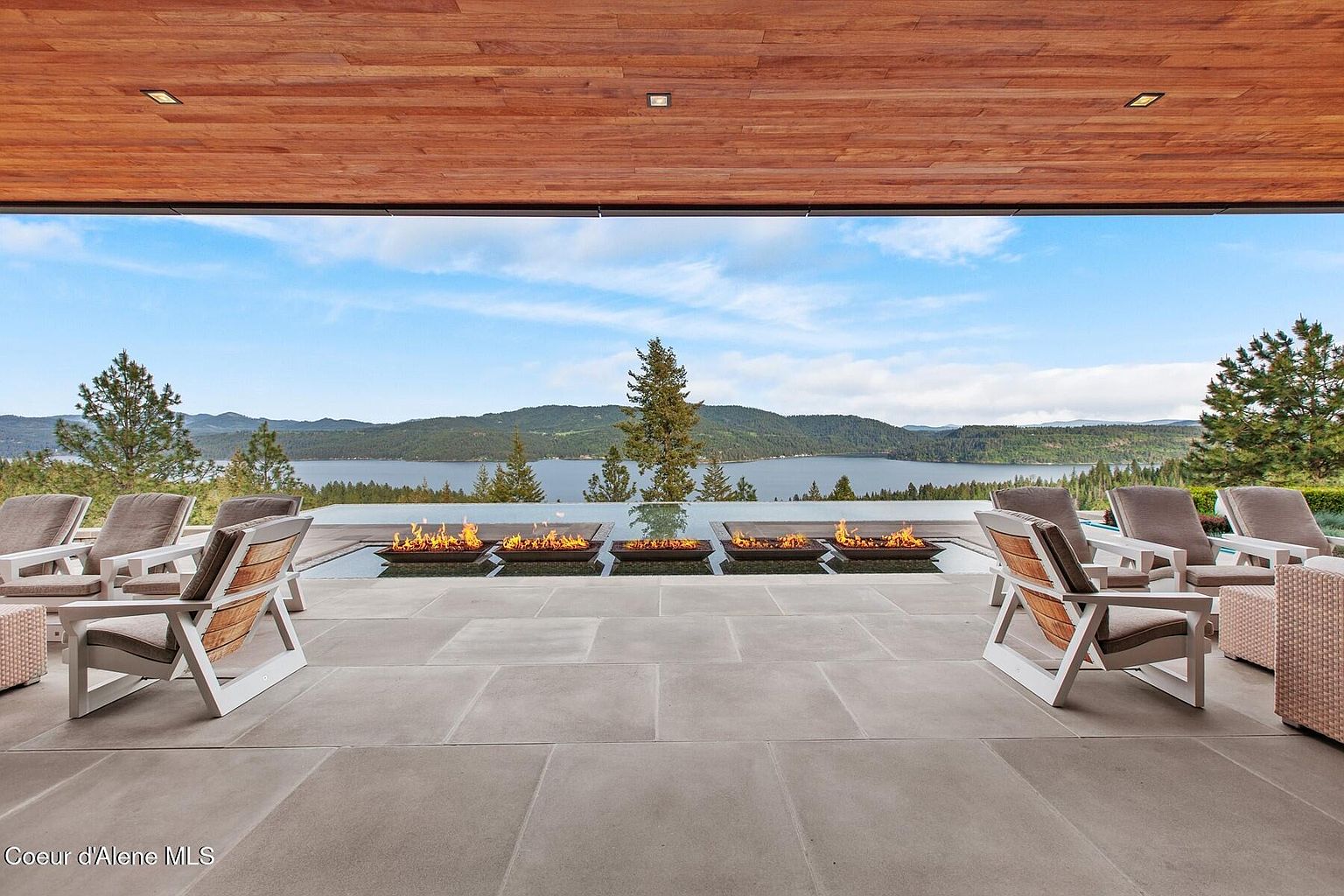 This is a stunning exterior shot of a patio or deck area, featuring modern outdoor furniture and a unique fire feature integrated into a water element. The space offers breathtaking views of a lake and mountains, creating a luxurious and serene atmosphere. The wooden ceiling adds a touch of warmth to the contemporary design.