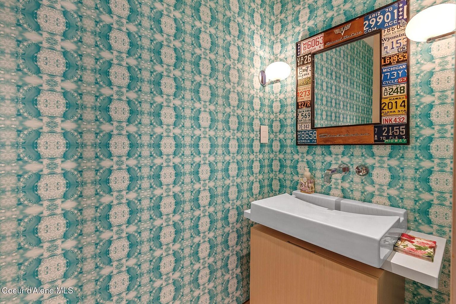 This is a quirky guest bathroom featuring a unique sink design with a built-in tissue holder and a mirror framed with vintage license plates. The walls are covered in a patterned wallpaper with a blue and white color scheme. The overall impression is eclectic and fun, making it a memorable space for guests.