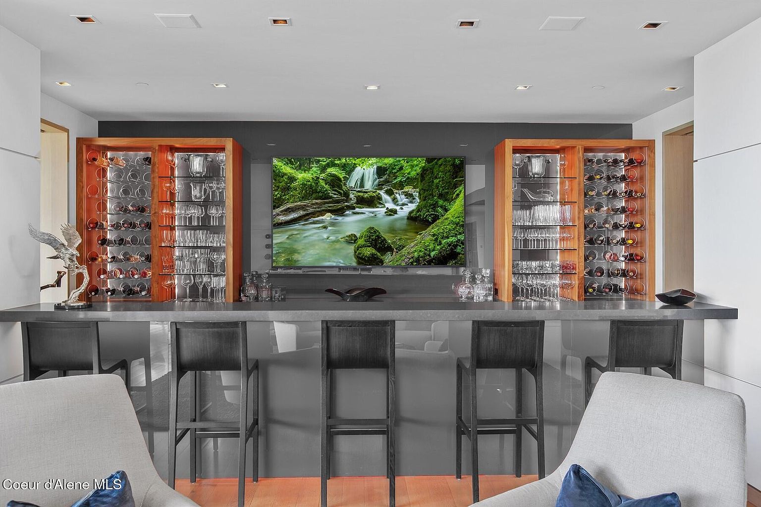 This image showcases a modern wine cellar or bar area, featuring custom wood cabinetry with glass doors displaying wine bottles and glassware. A sleek, gray countertop serves as a bar, complemented by modern bar stools. A large screen displays a nature scene, adding a unique focal point to the space, and the overall design is sophisticated and inviting.