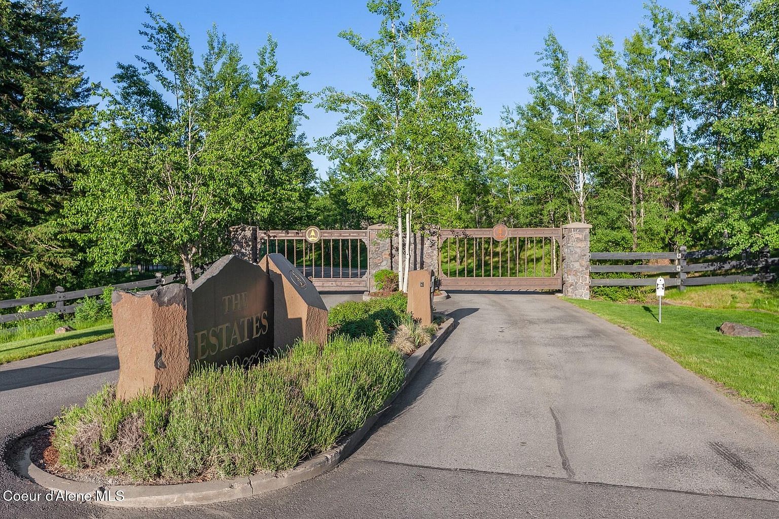 The image showcases the grand entryway to "The Estates," featuring a stone sign with the community's name, flanked by lush greenery. An ornate metal gate, set within stone pillars, marks the entrance, with mature trees lining the background. The paved driveway leads into the property, suggesting exclusivity and well-maintained grounds.