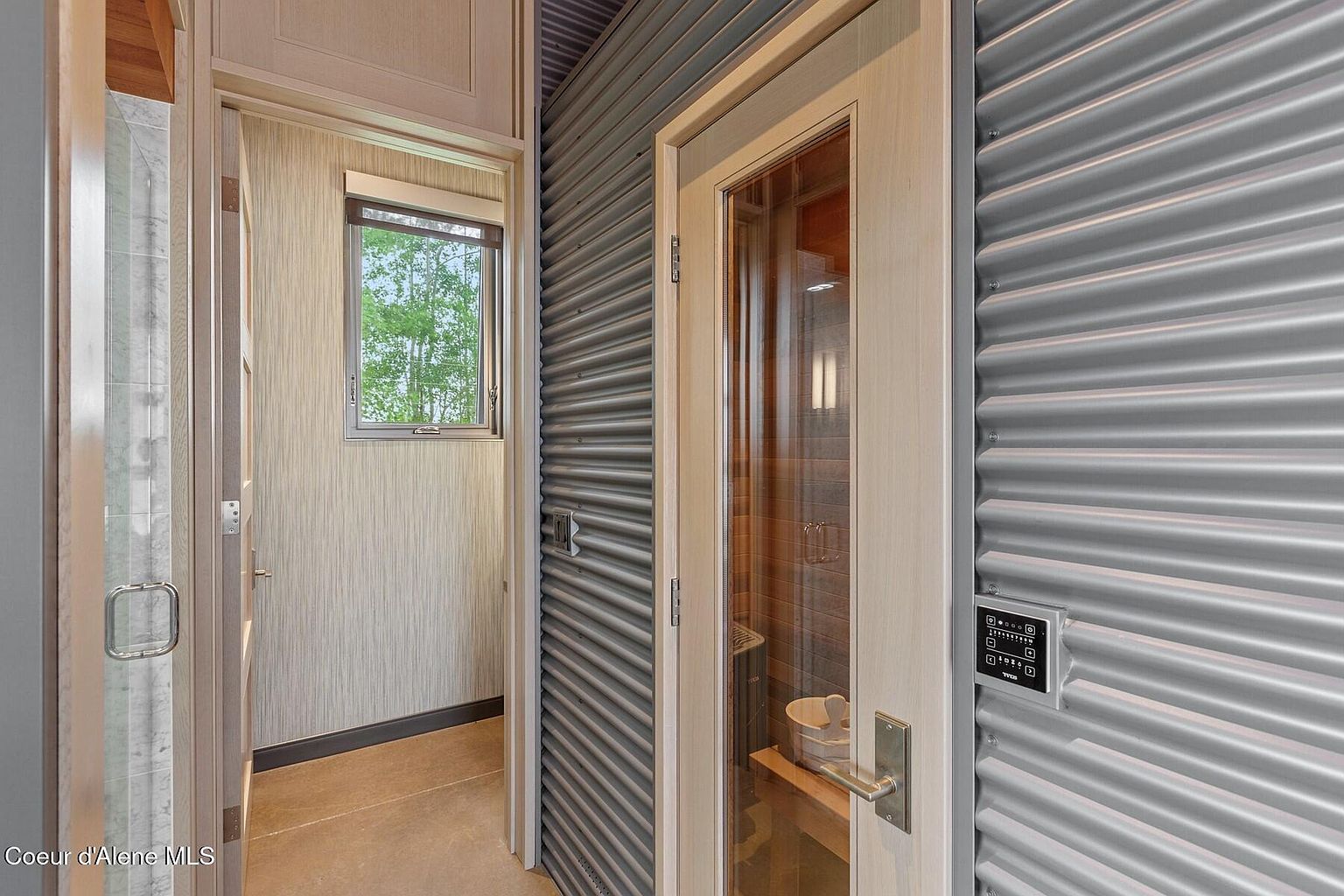 The image showcases a modern bathroom interior, featuring a sauna with a glass door and a control panel mounted on the corrugated metal wall. A doorway leads to another section of the bathroom with a window providing natural light. The design incorporates a blend of textures and materials, creating a spa-like atmosphere.