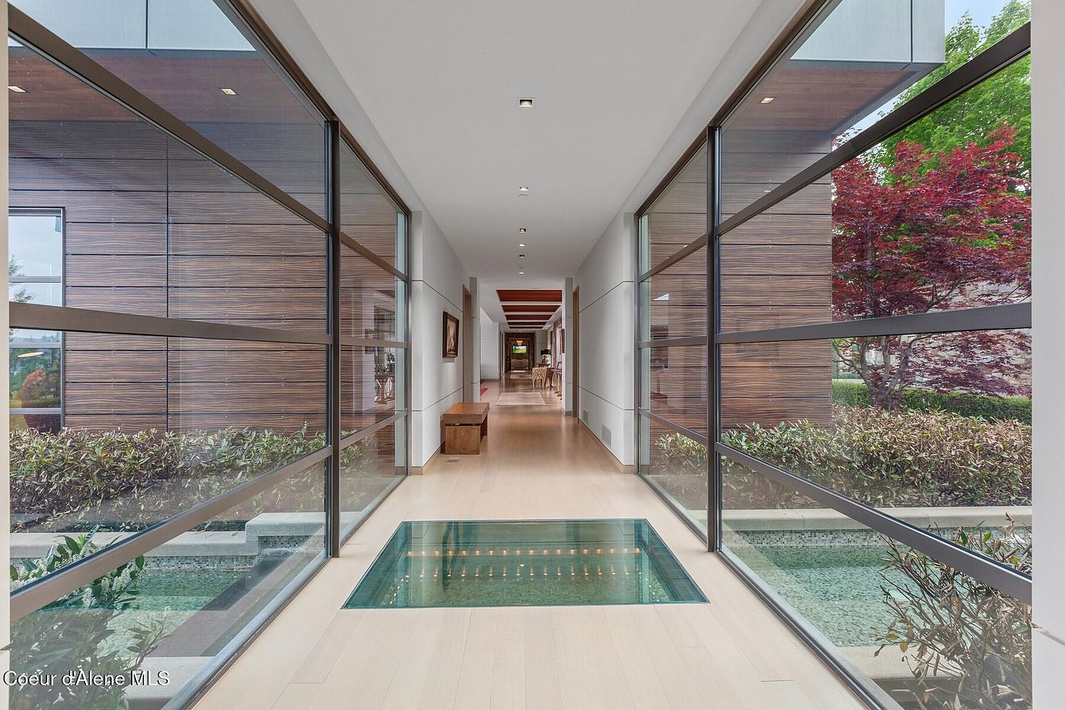 This is an interior shot of a modern hallway featuring a unique glass-covered water feature in the floor. The hallway is lined with large windows offering views of the exterior landscaping, and the walls are painted in a neutral tone. The flooring is light wood, and the overall design is sleek and contemporary.