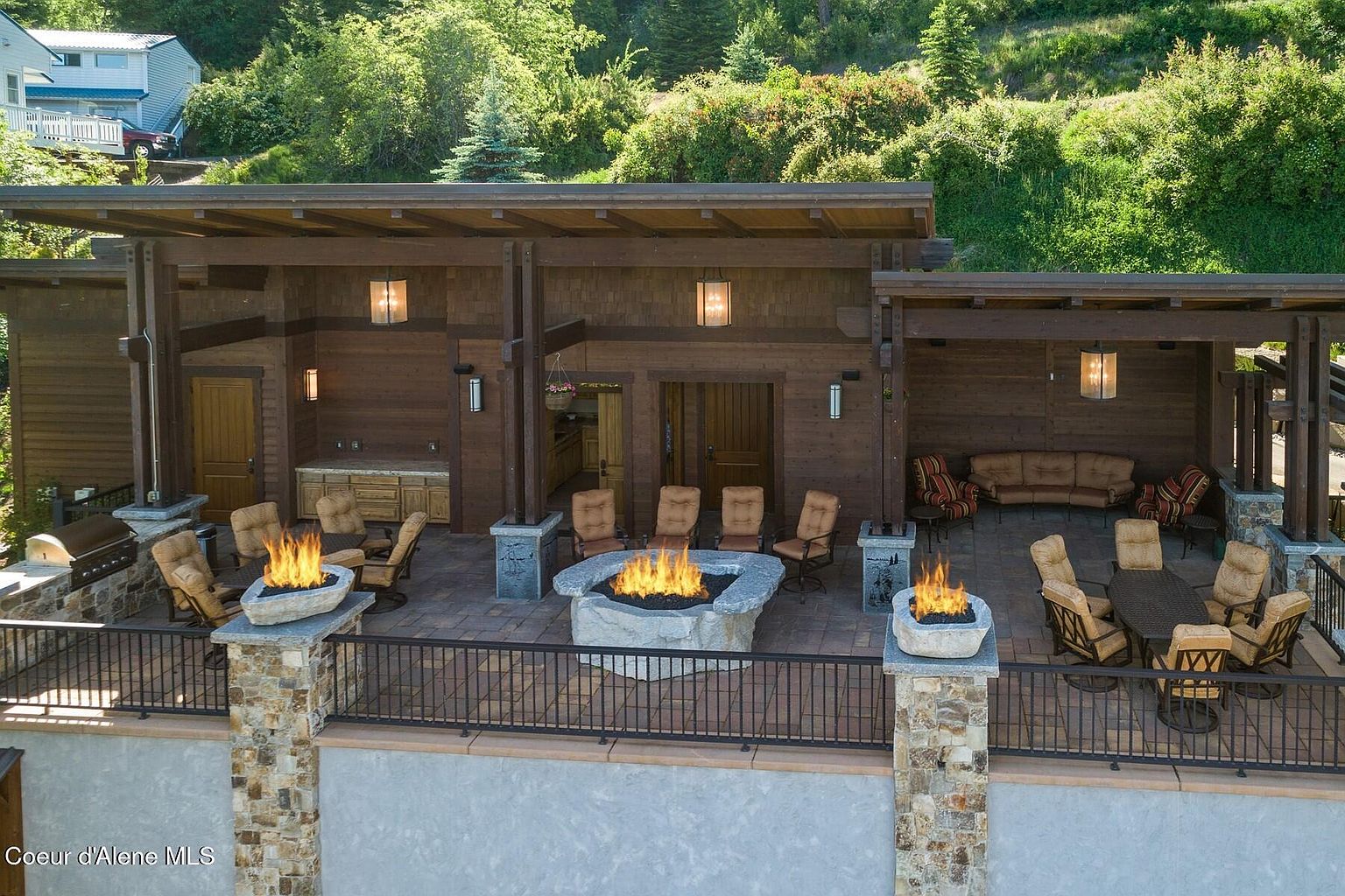 This image showcases a luxurious outdoor living space, featuring a covered patio with wooden beams and siding. The patio includes a built-in grill area, comfortable seating around a stone fire pit, and a dining area with a table and chairs. The space is elevated with a stone and metal railing, offering views of the surrounding greenery and creating an inviting atmosphere for relaxation and entertainment.