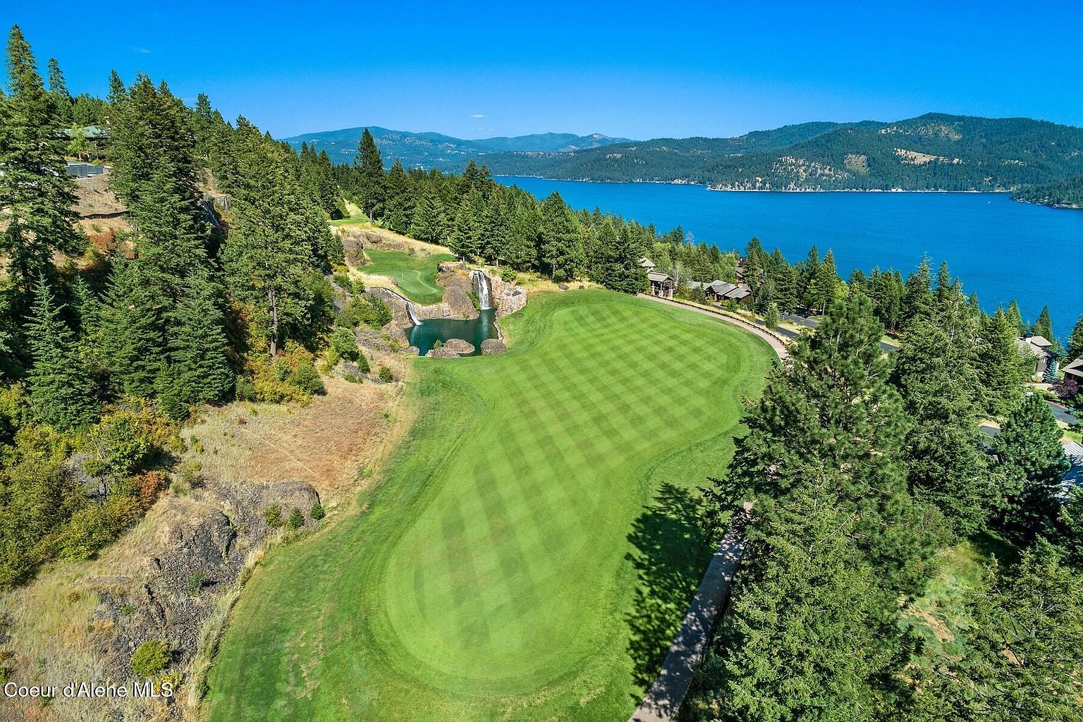 This aerial shot showcases a beautifully manicured golf course green with a unique water feature, including a waterfall and pond. The green is surrounded by lush trees and overlooks a stunning lake with mountains in the distance, creating a picturesque and luxurious setting. The image highlights the property's natural beauty and recreational amenities.