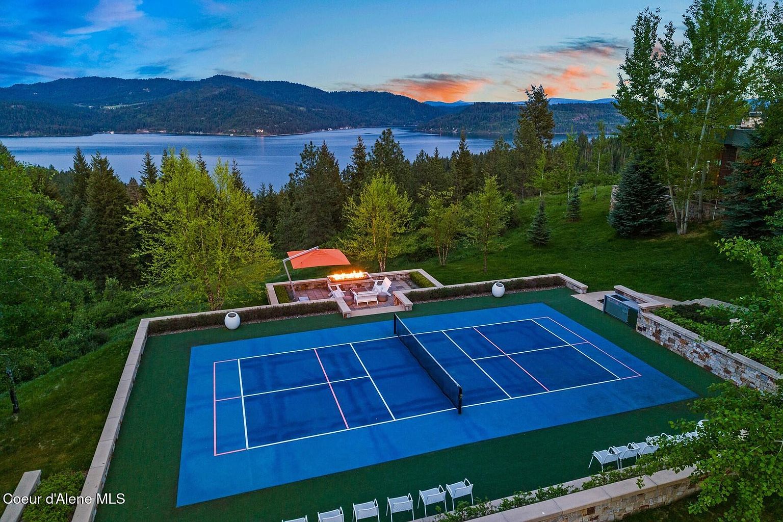 This aerial shot showcases a stunning property featuring a blue tennis court with green surroundings, a cozy fire pit area with seating, and a breathtaking view of a lake and mountains in the background. The well-maintained landscaping and the overall layout suggest a luxurious and private estate, perfect for outdoor recreation and relaxation. The image is taken from a high angle, providing a comprehensive view of the property's layout and its scenic surroundings.