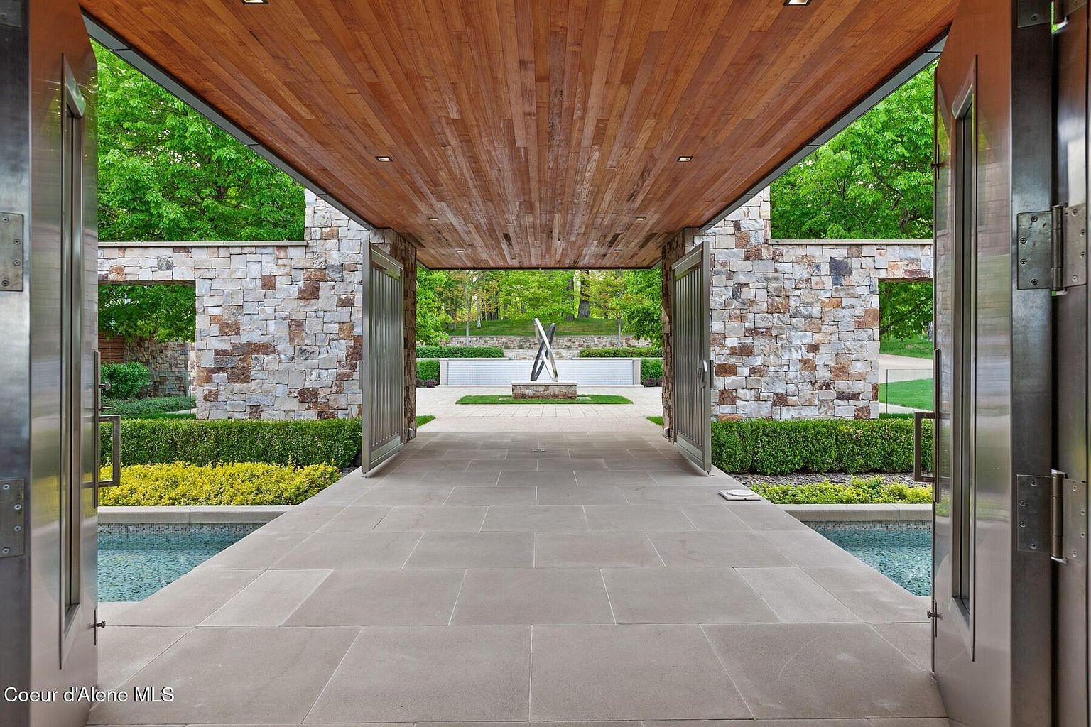 This image showcases a grand entryway featuring a wooden ceiling, stone walls, and large metal gates that open to a beautifully landscaped garden. The pathway is paved with large stone tiles, leading to a modern sculpture and a serene water feature. The overall impression is one of luxury and sophisticated design, creating an inviting and impressive entrance to the property.