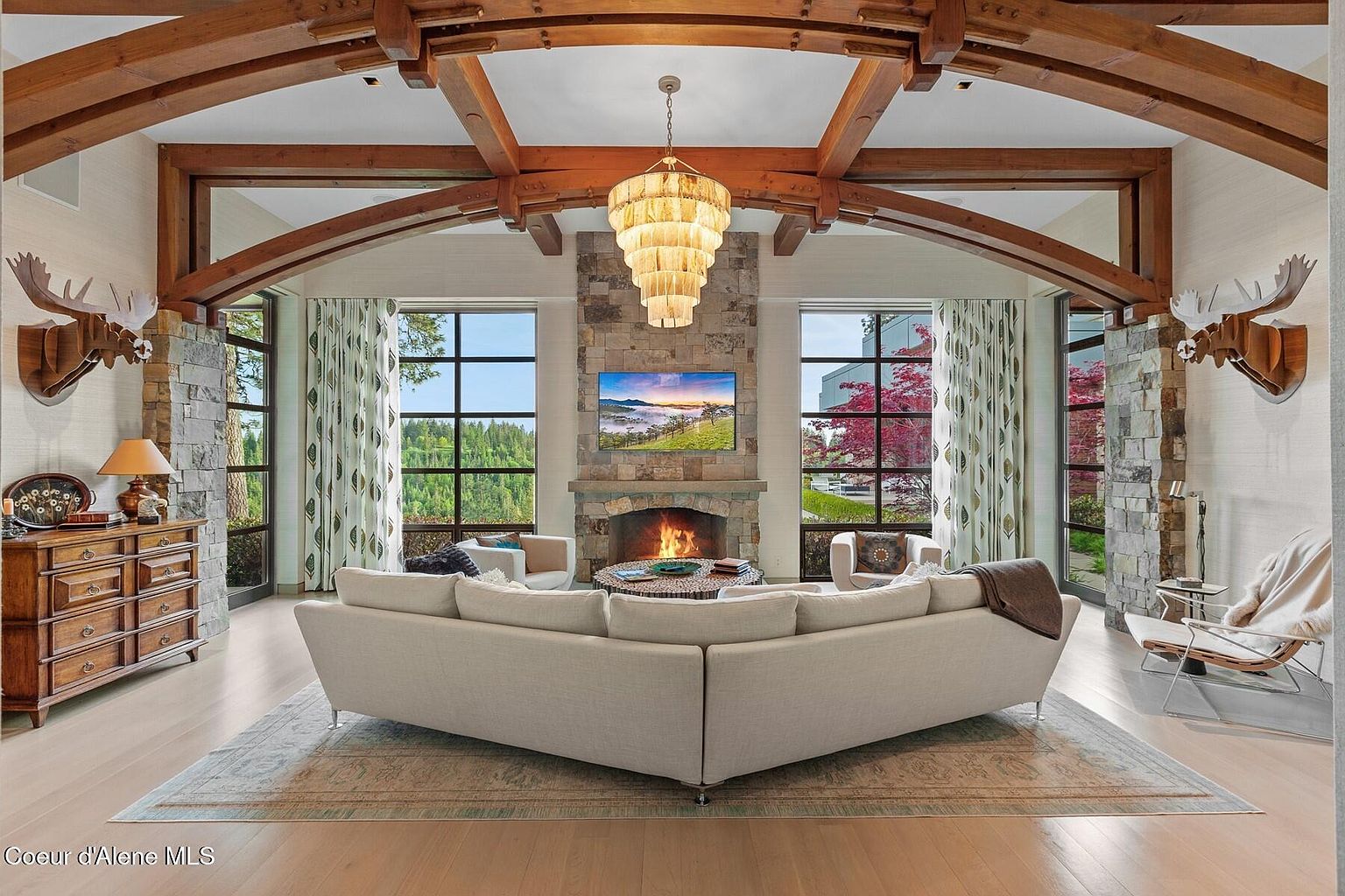 This is an interior shot of a luxurious living room featuring a stone fireplace with a mounted television, flanked by large windows offering views of the surrounding landscape. The room is furnished with a large sectional sofa, a unique chandelier, and wooden architectural beams, creating a warm and inviting atmosphere. The decor includes wooden moose head wall decor and a wooden dresser.