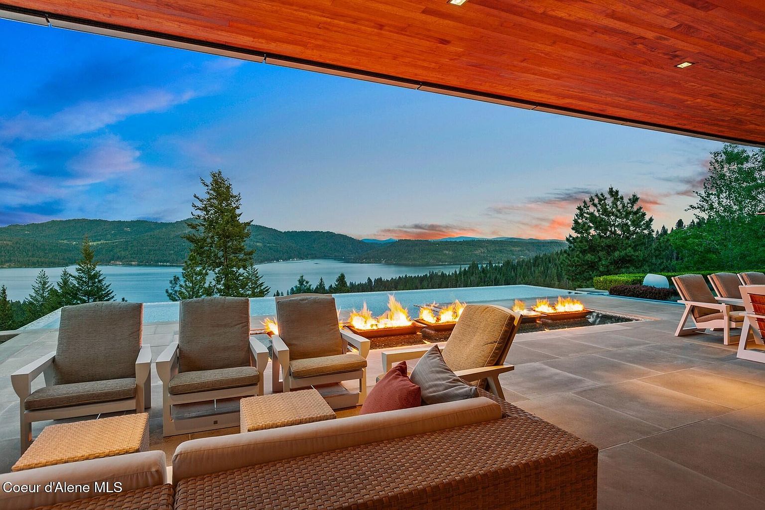 This image showcases a luxurious outdoor patio or deck area with stunning lake views. The space features comfortable seating arrangements, including chairs and a wicker sofa, complemented by small tables. A modern fire pit adds warmth and ambiance, while the infinity pool seamlessly blends with the lake horizon, creating a serene and inviting atmosphere.