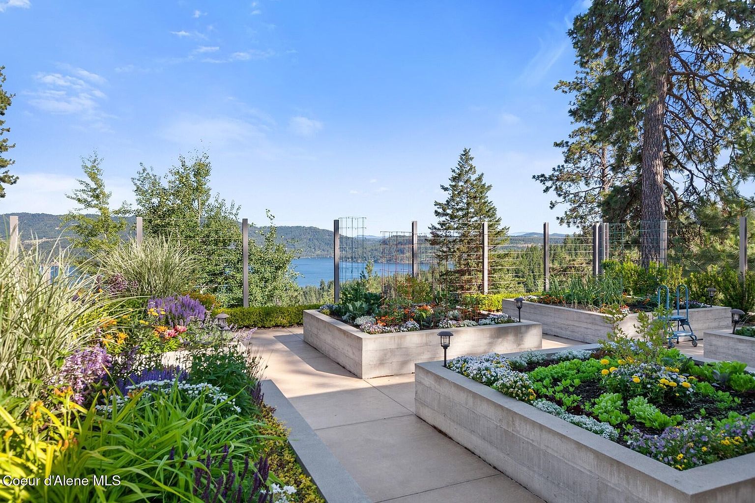 This image showcases a beautifully landscaped garden with raised concrete planters filled with a variety of flowers and plants. The garden overlooks a serene lake and distant mountains, creating a picturesque and tranquil setting. A wire fence provides a subtle boundary without obstructing the stunning view.